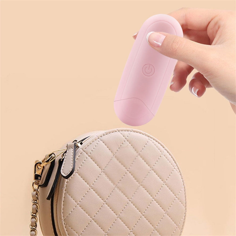 Hand hält rosa Gerät über einer Handtasche. Beiger Hintergrund. Power-Taste sichtbar.