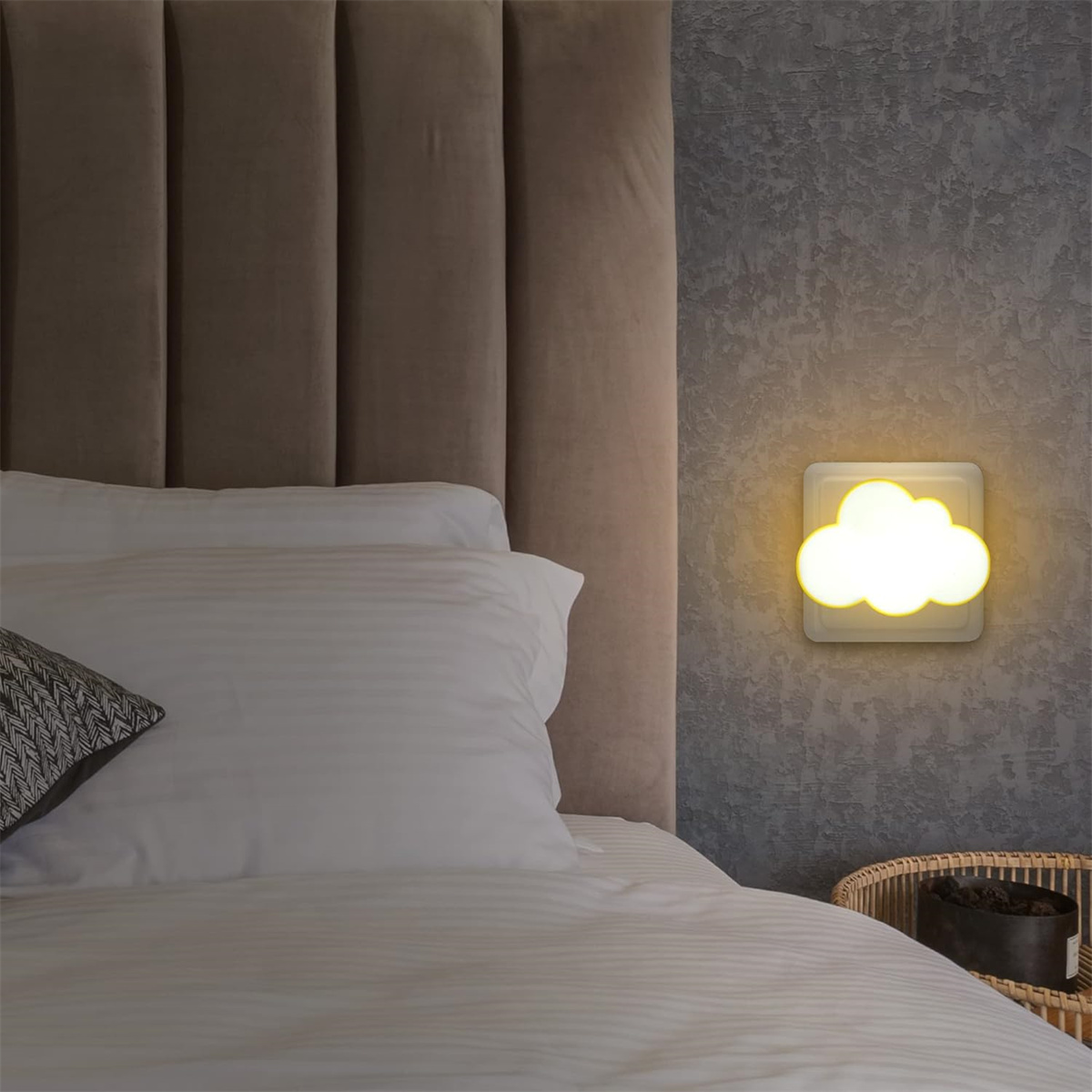 Eine wolkenförmige Wandlampe, die gelbes Licht abgibt. Sie befindet sich an einer grauen Wand über einem Bett.