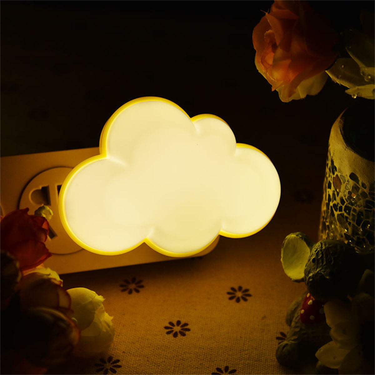 Ein wolkenförmiges Nachtlicht leuchtet gelb, neben einem hölzernen Stecker. Im Hintergrund eine Vase mit Blumen.