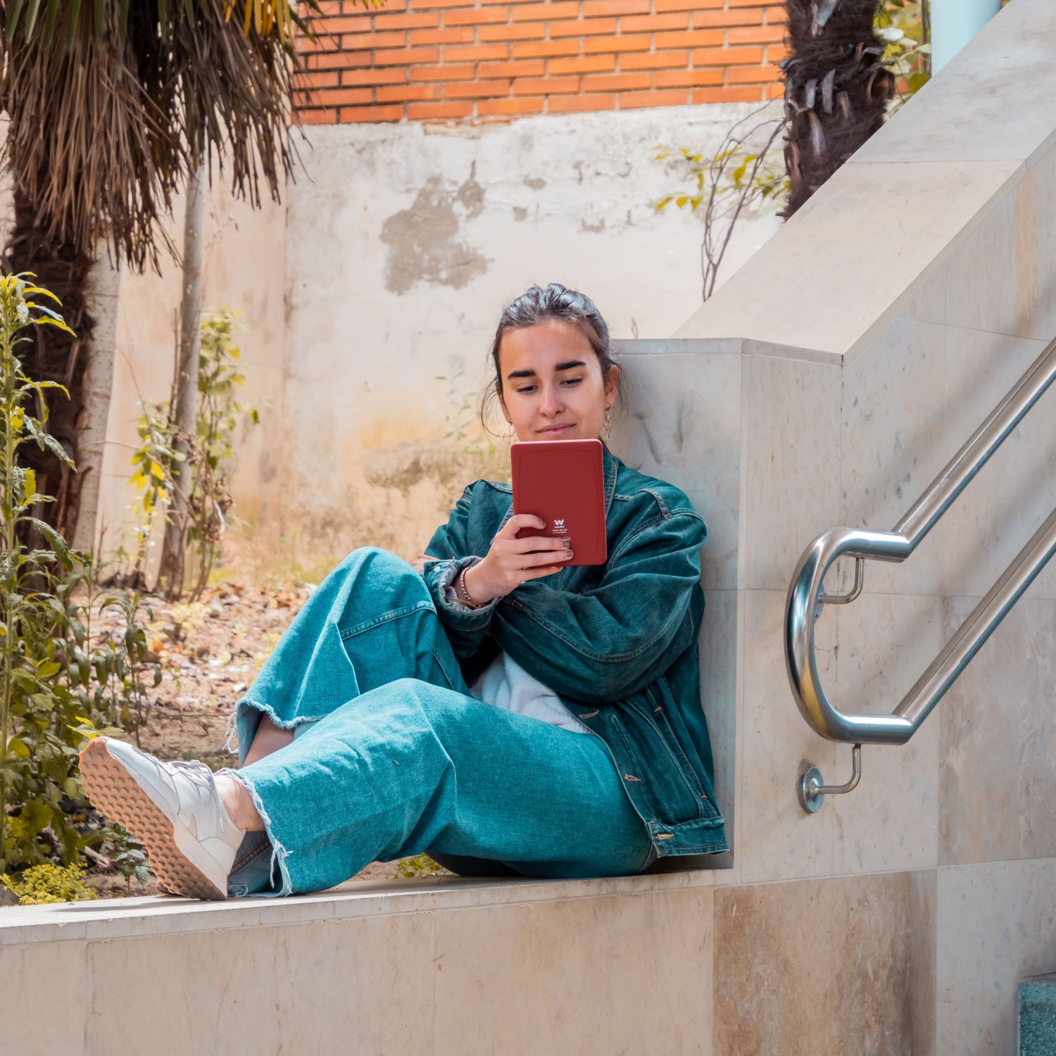 Kobieta siedzi, czyta czerwony czytnik e-booków, w dżinsowej kurtce i dżinsach.