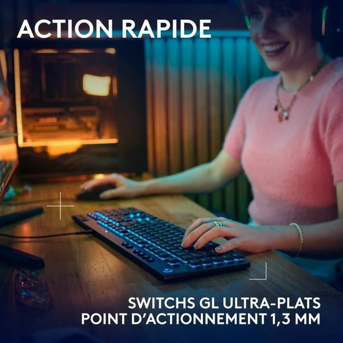 Kobieta gra w grę z klawiaturą, myszą i monitorem. Klawiatura ma niebieskie światła. Drewniane biurko.