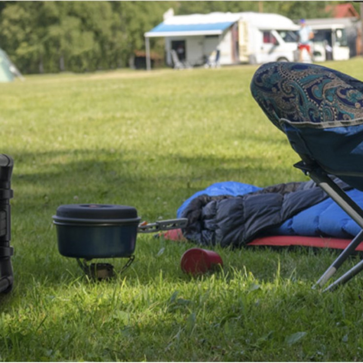 Scena kempingowa z krzesłem, garnkiem, śpiworem, czerwonym kubkiem na trawie; tło drzew i budynków.