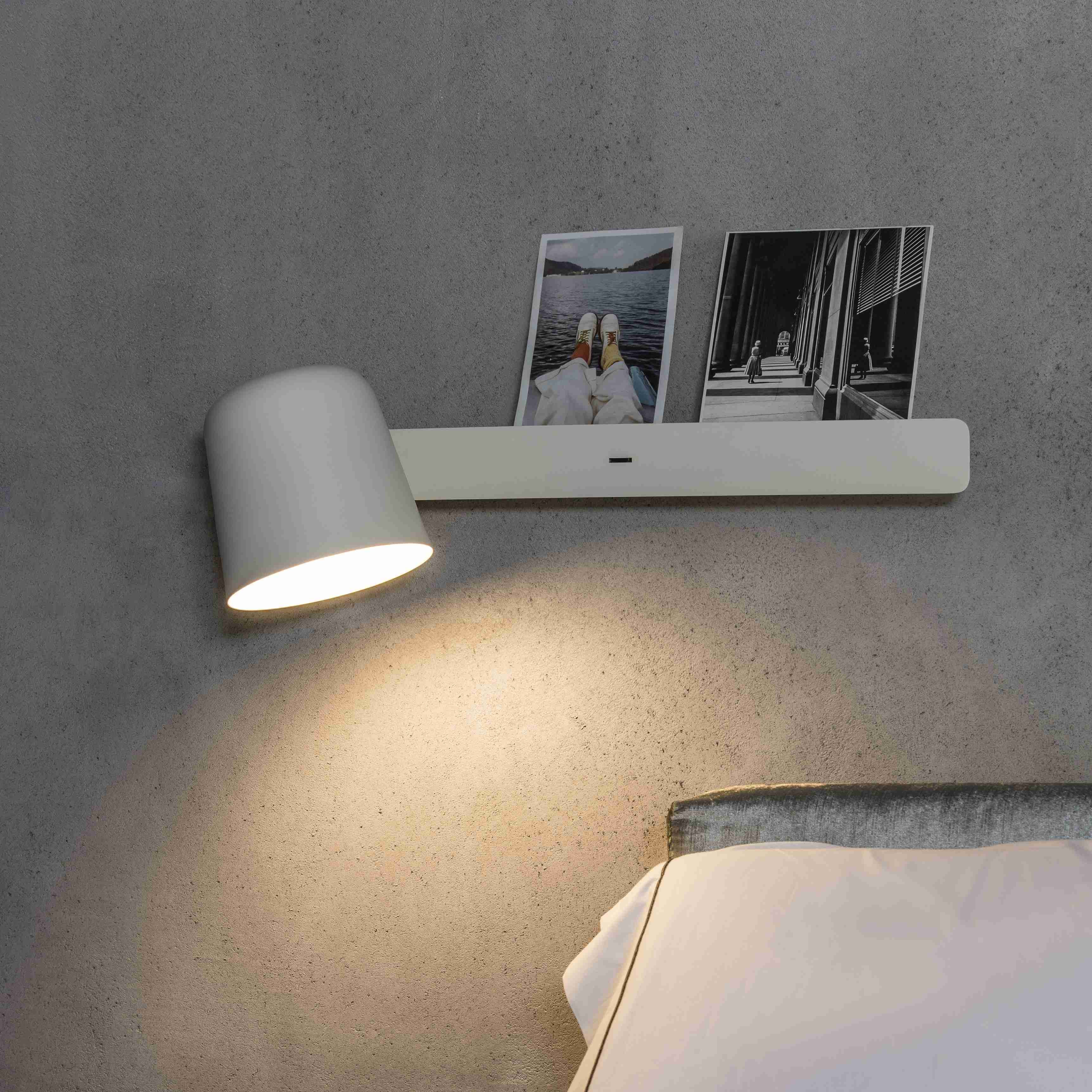 Lampada da parete con paralume bianco e mensola con foto, che proietta la luce verso il basso.