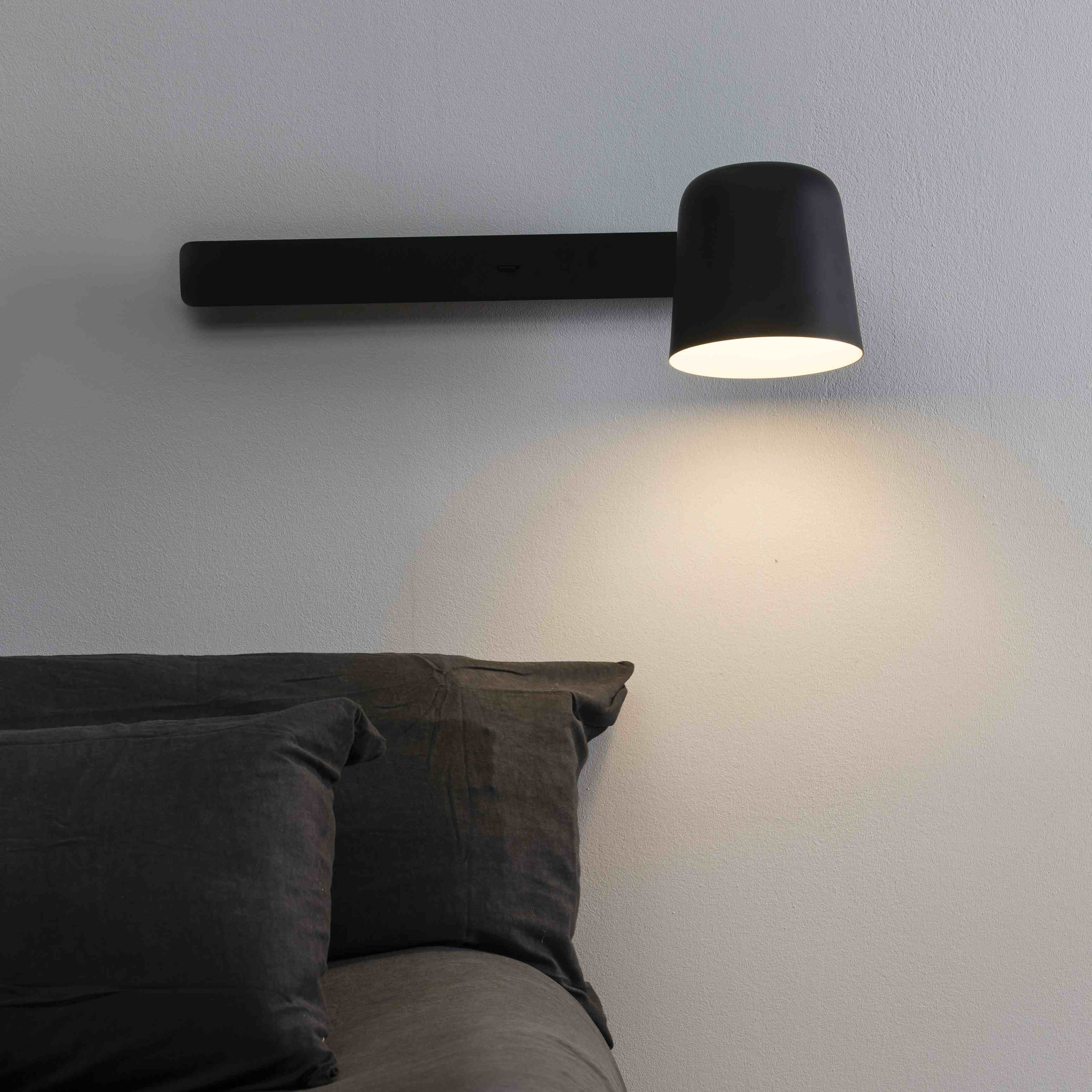 Lampada da parete nera con paralume cilindrico, montata su una parete grigia sopra un cuscino grigio.