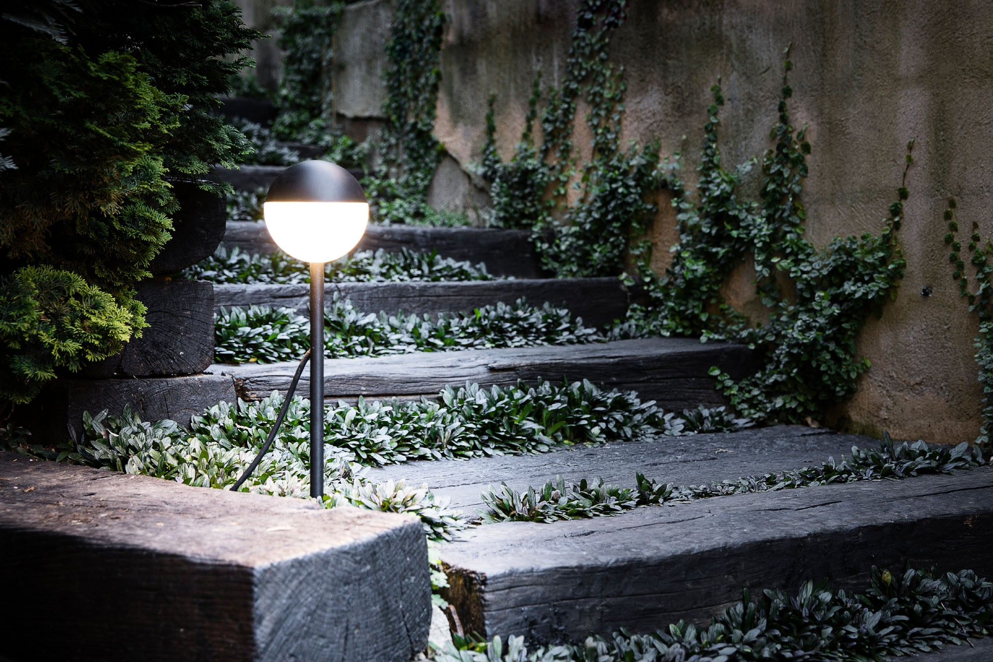 Una lampada rotonda e nera su un palo illumina gradini di legno in un giardino.