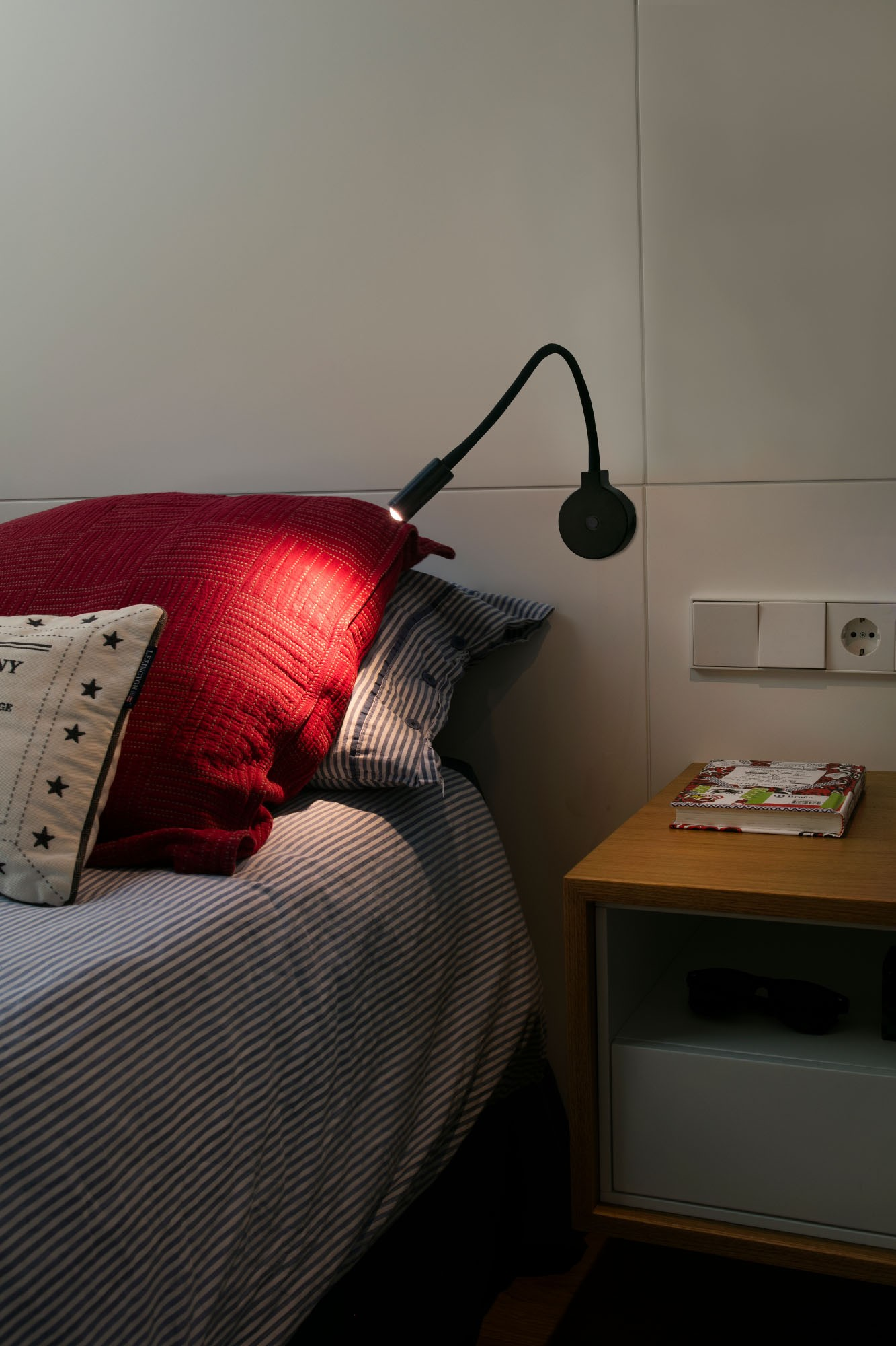 Letto con cuscino rosso, lenzuola a righe. Lampada da parete nera vicino al comodino.