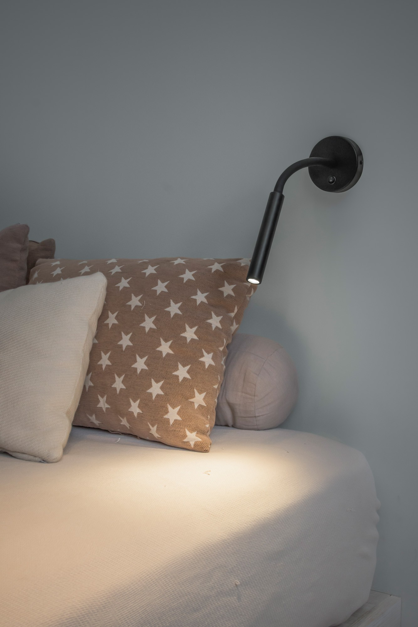 Lampada da parete per comodino con braccio nero e luce accesa, accanto a cuscini e letto.
