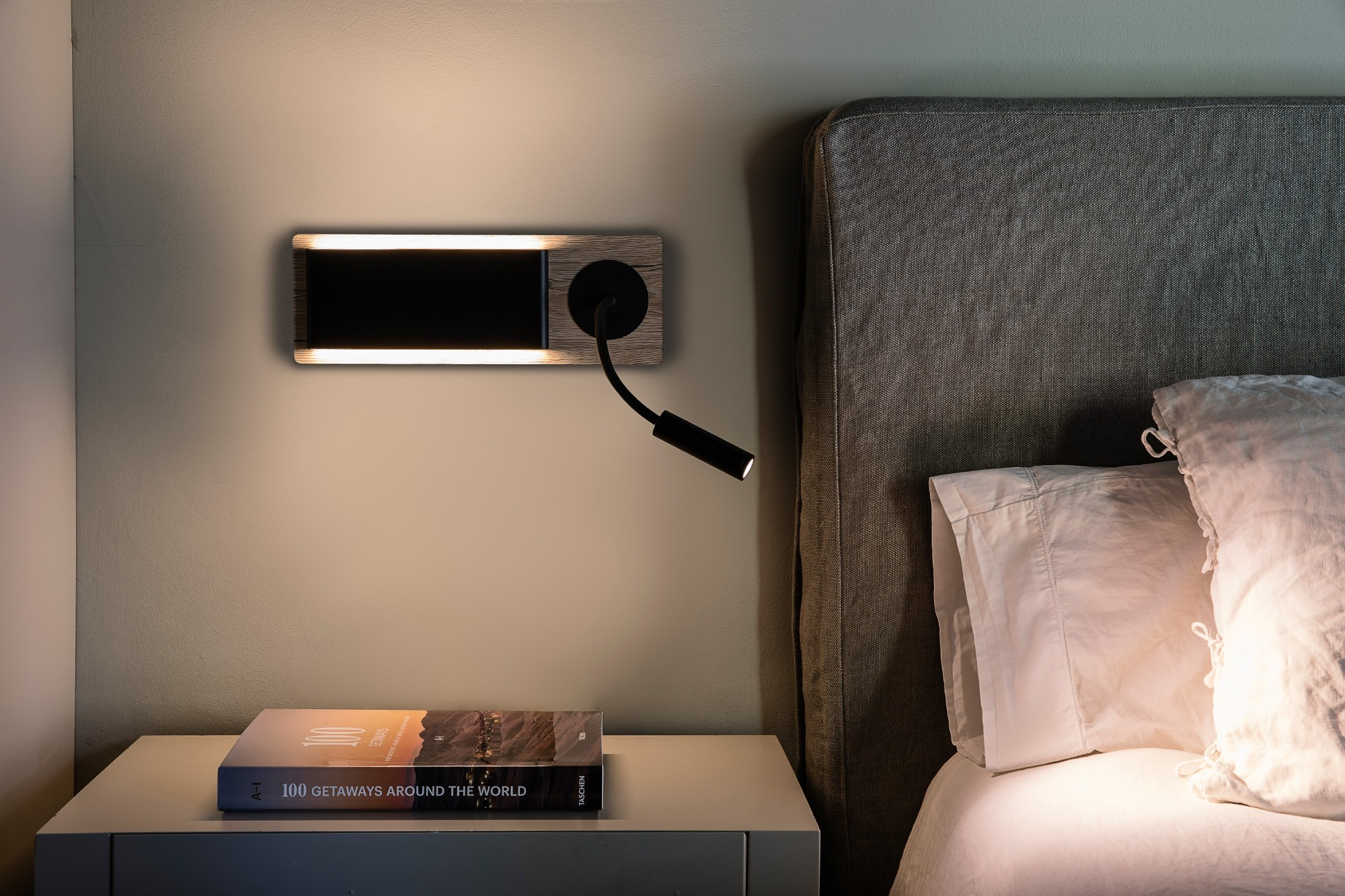 Una scena del comodino: una lampada da parete, un libro e parte di un letto. La luce calda è accesa.
