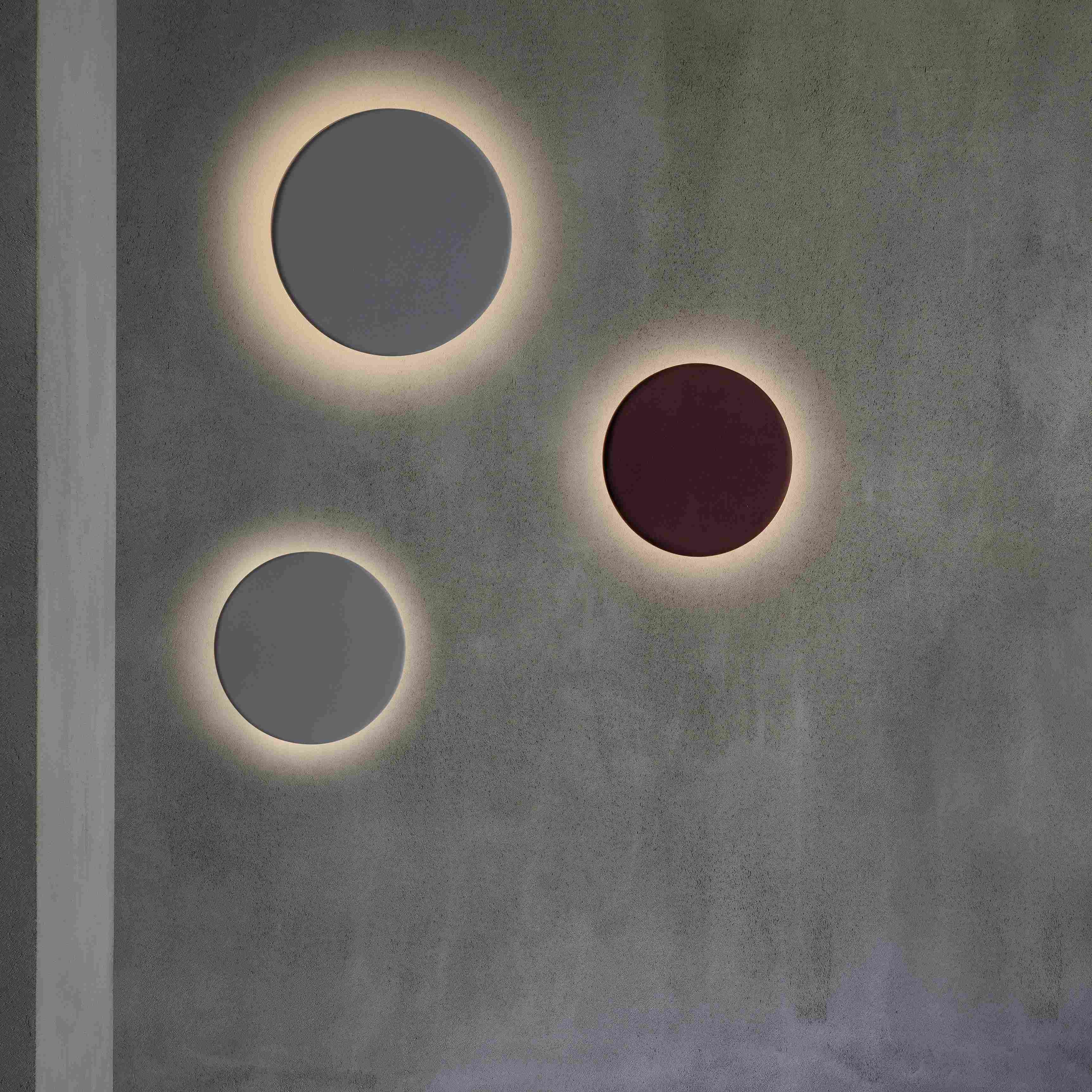 Tre lampade da parete circolari su un muro grigio. Quella centrale è rossa, le altre grigie.