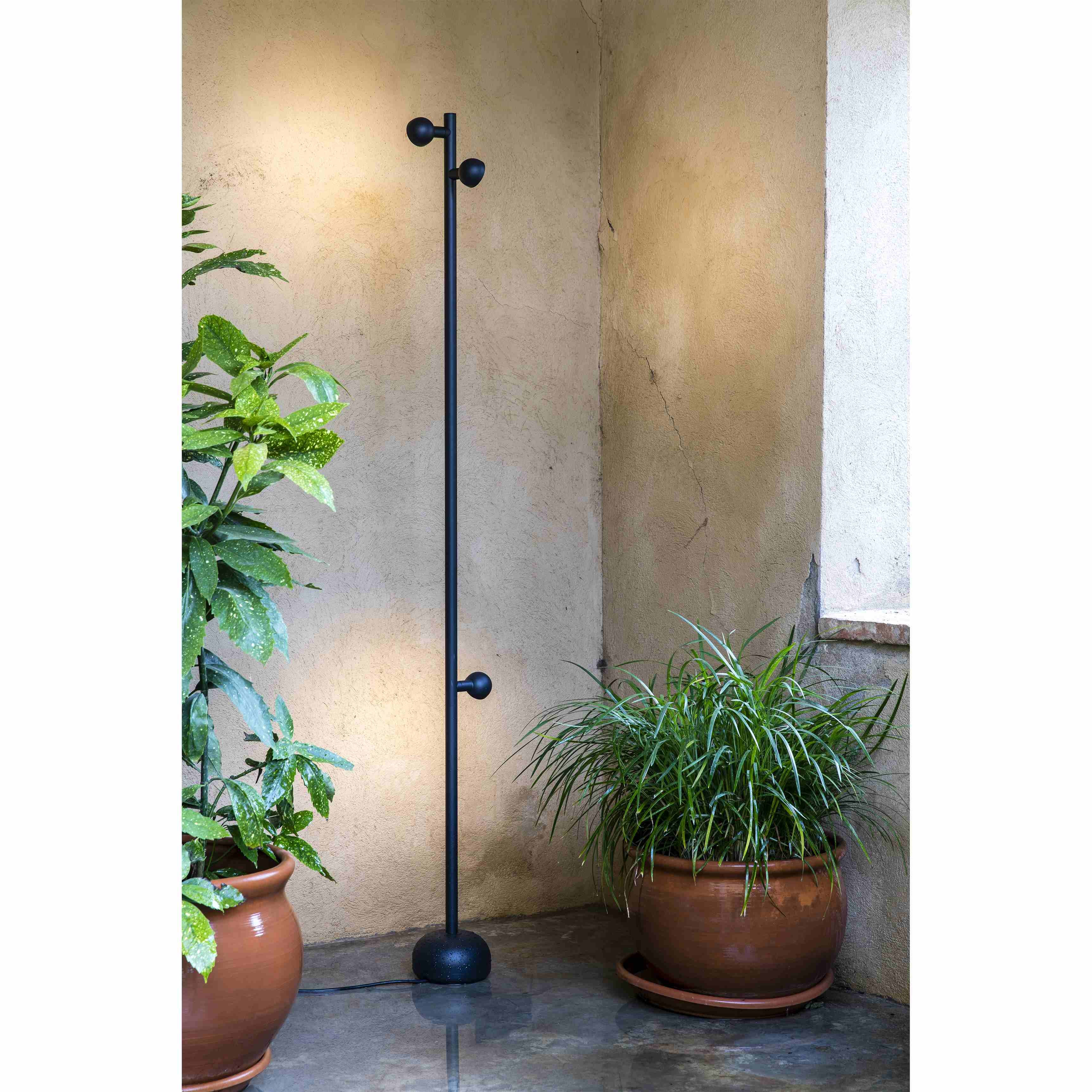 Un'apparecchio di illuminazione per esterni nero con tre luci su un palo alto, vicino a piante in vaso.