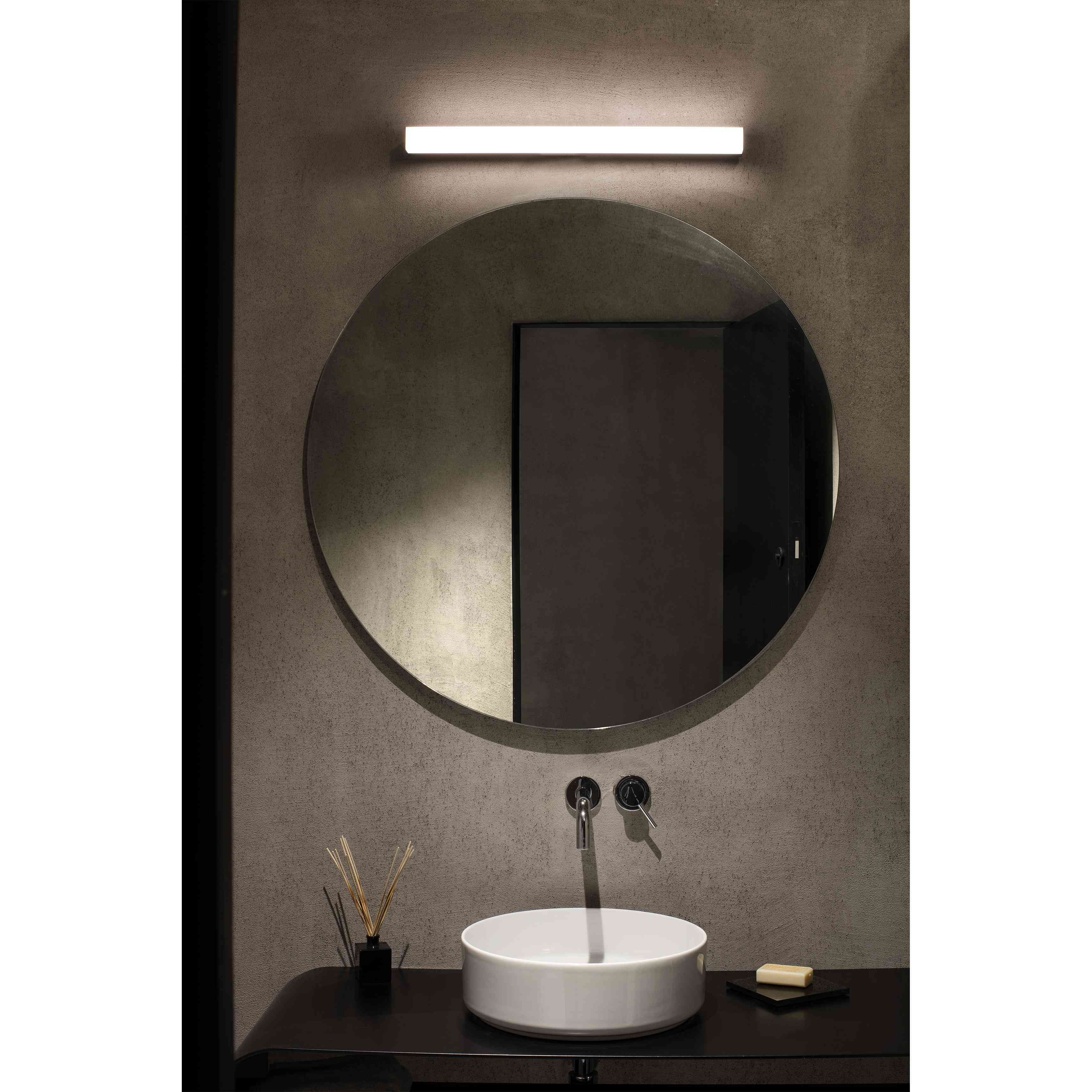 Bagno con specchio rotondo sopra un lavandino, una piccola lampada e un vassoio di bastoncini profumati. Parete grigia.