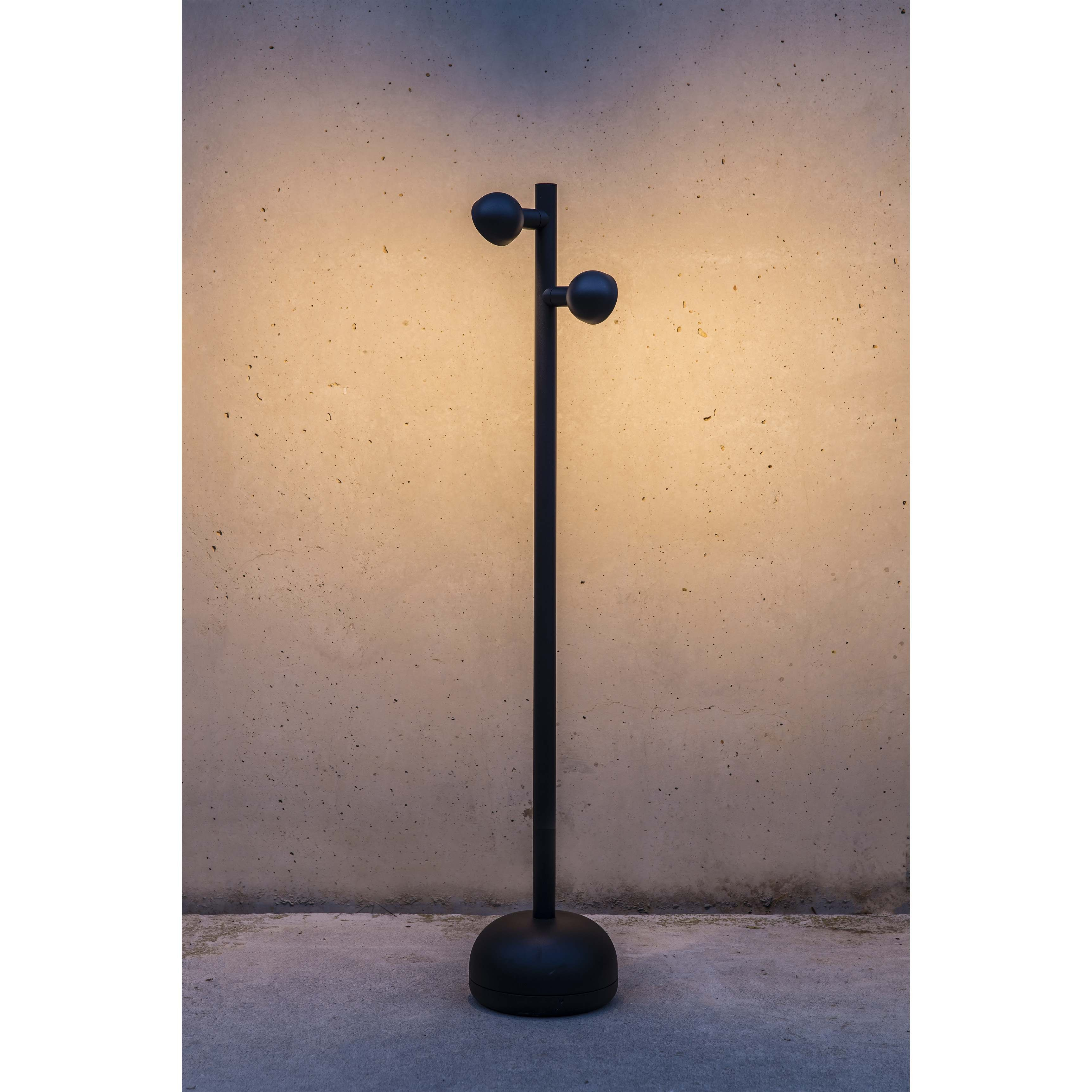 Lampada da esterno nera su un palo. Ha due luci. Su una superficie di cemento. Sfondo grigio.