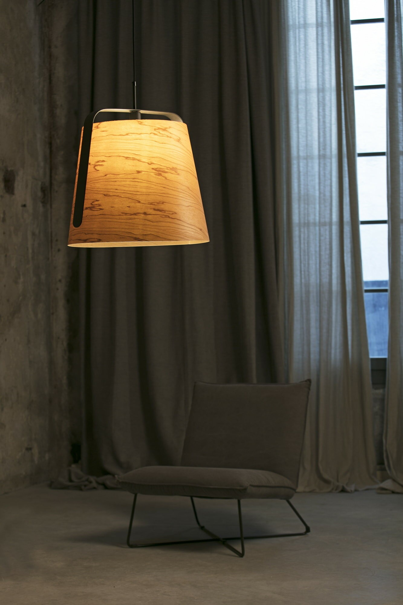 Un paralume con venature del legno pende dal soffitto, con una sedia in primo piano. Muro di cemento grigio e tende.