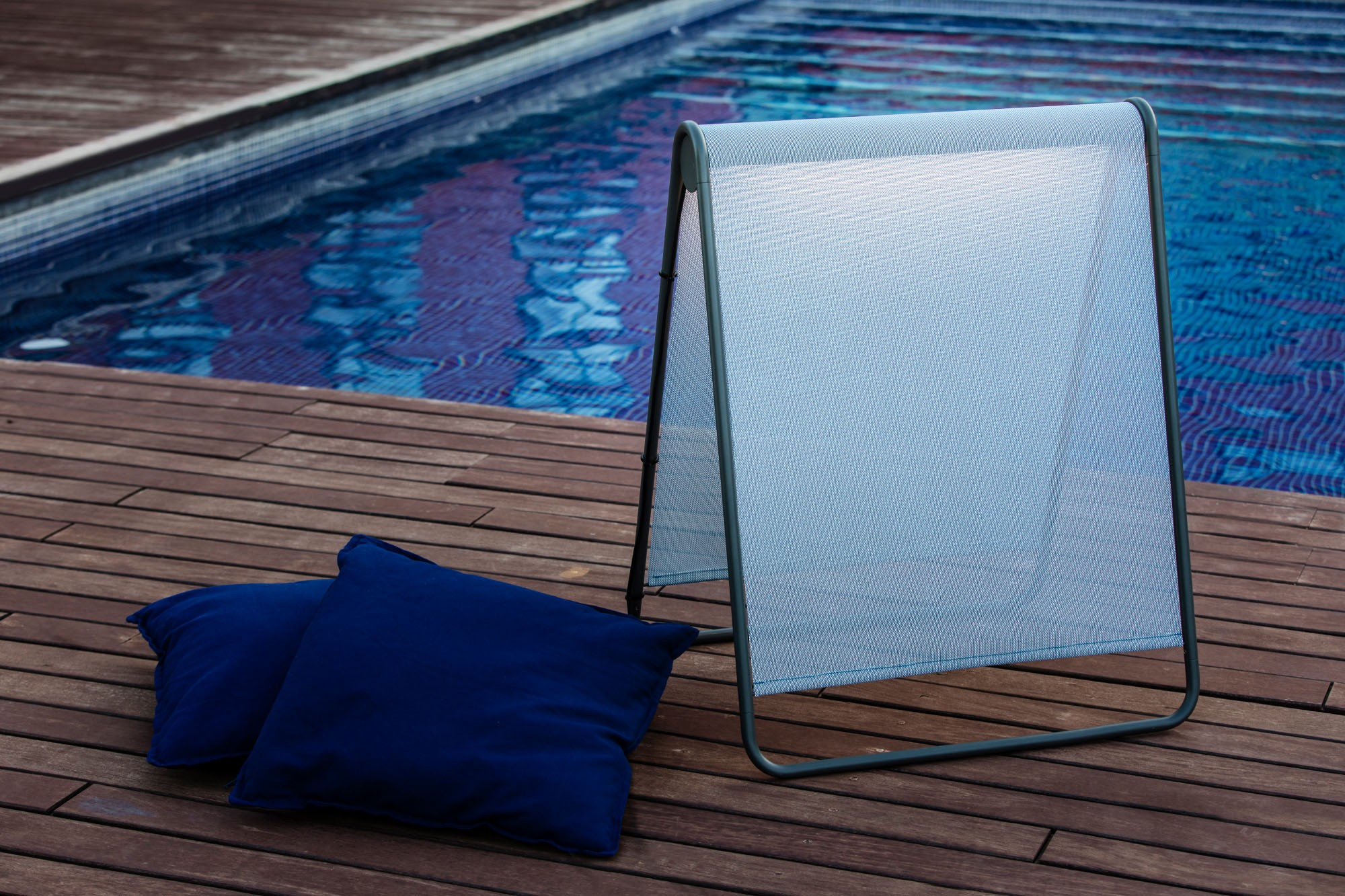 Parasole a forma di A vicino a una piscina. Due cuscini blu sono sul ponte di legno.