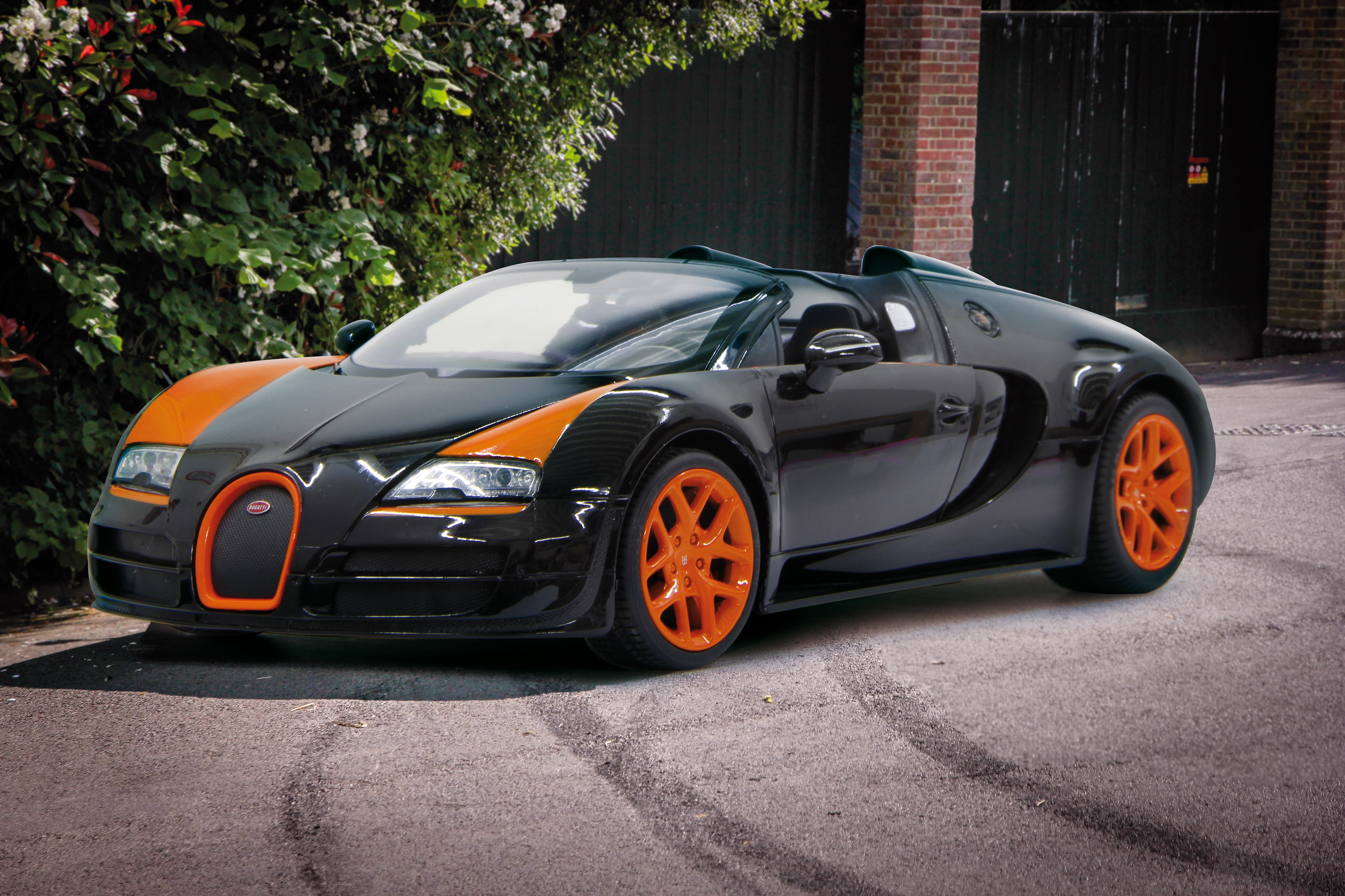 Czarny i pomarańczowy samochód sportowy zaparkowany na ulicy. Zielone krzewy i ceglany budynek w tle.