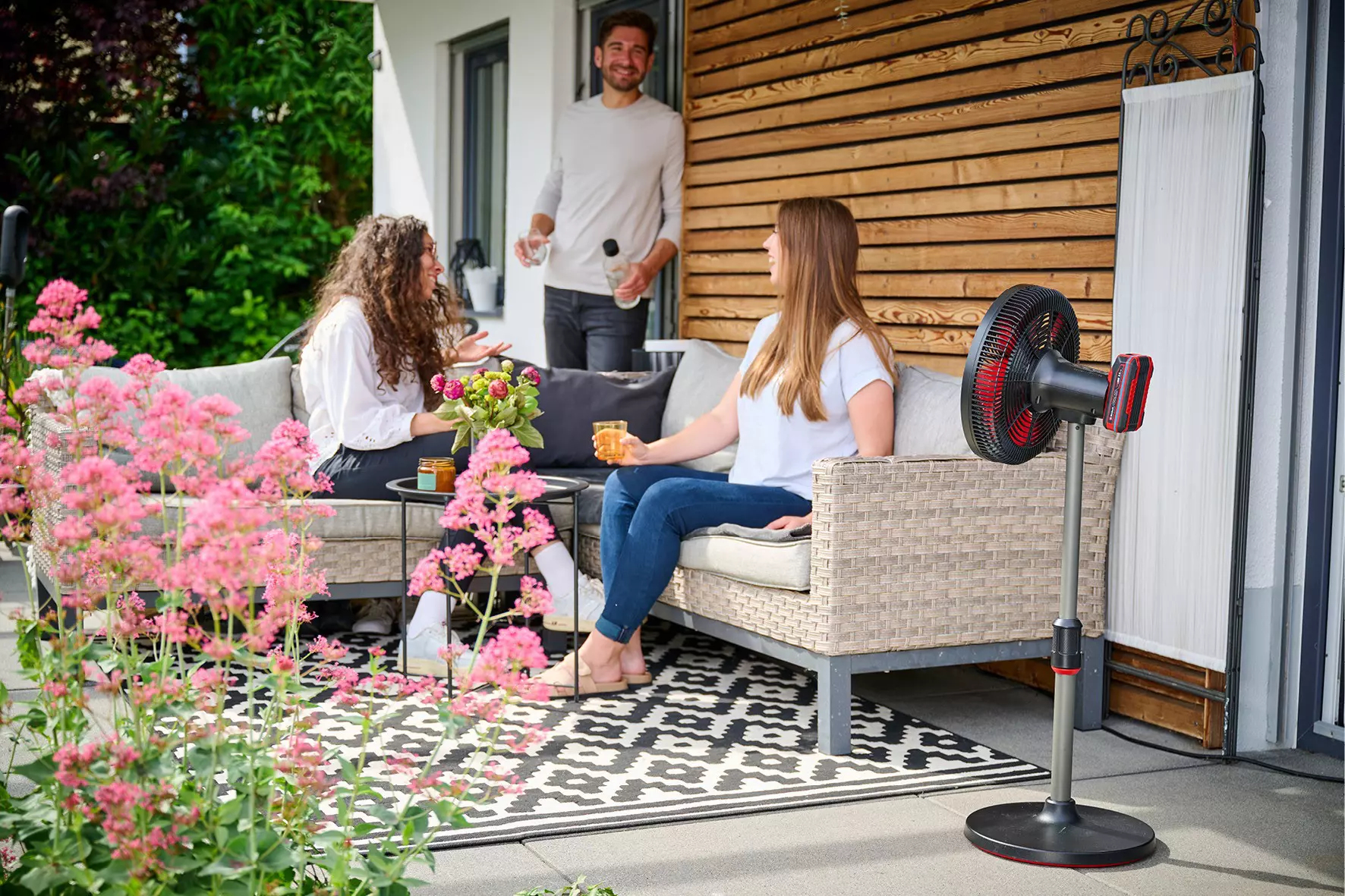 Dwie kobiety i mężczyzna na patio z wentylatorem, różowymi kwiatami i drewnianą ścianą.