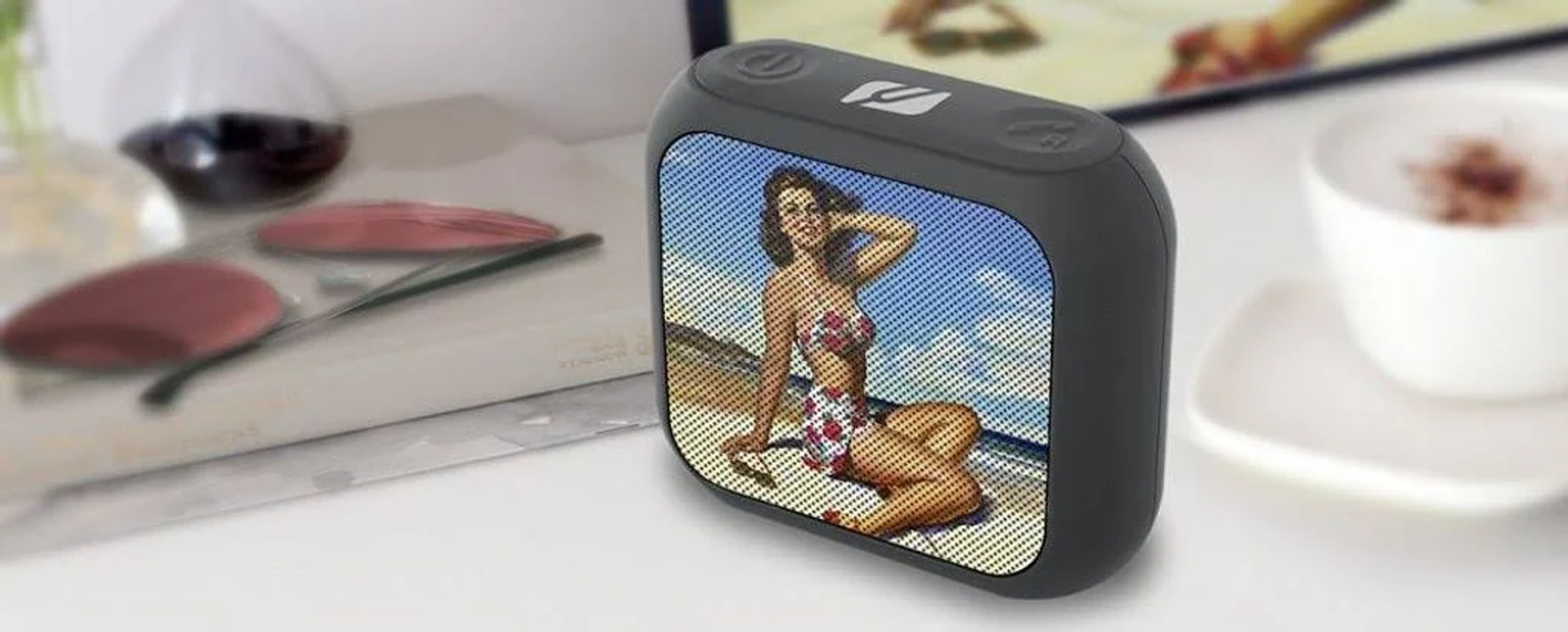 Un petit haut-parleur gris foncé montre une pin-up et est posé sur une surface blanche.