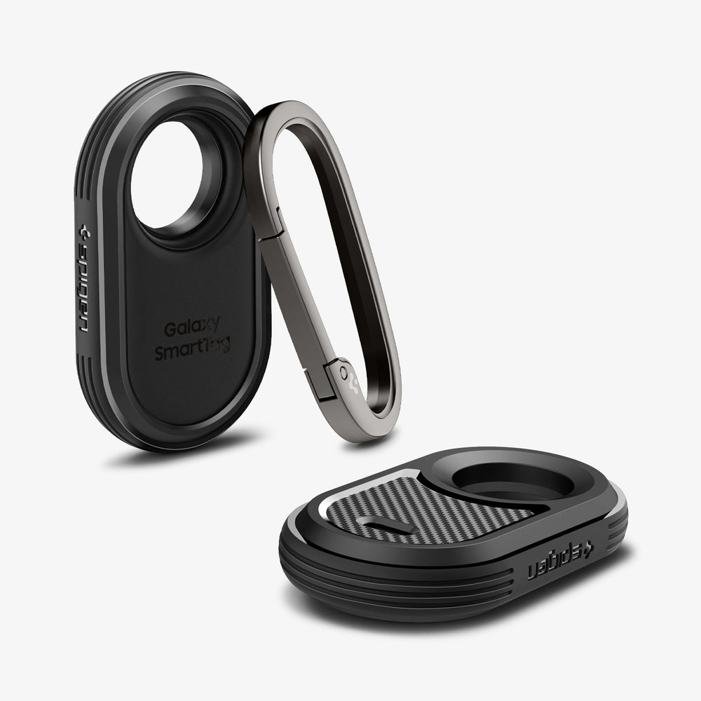 Dwa czarne uchwyty Spigen smart tag z otwartym srebrnym karabińczykiem, na jasnoszarej powierzchni.