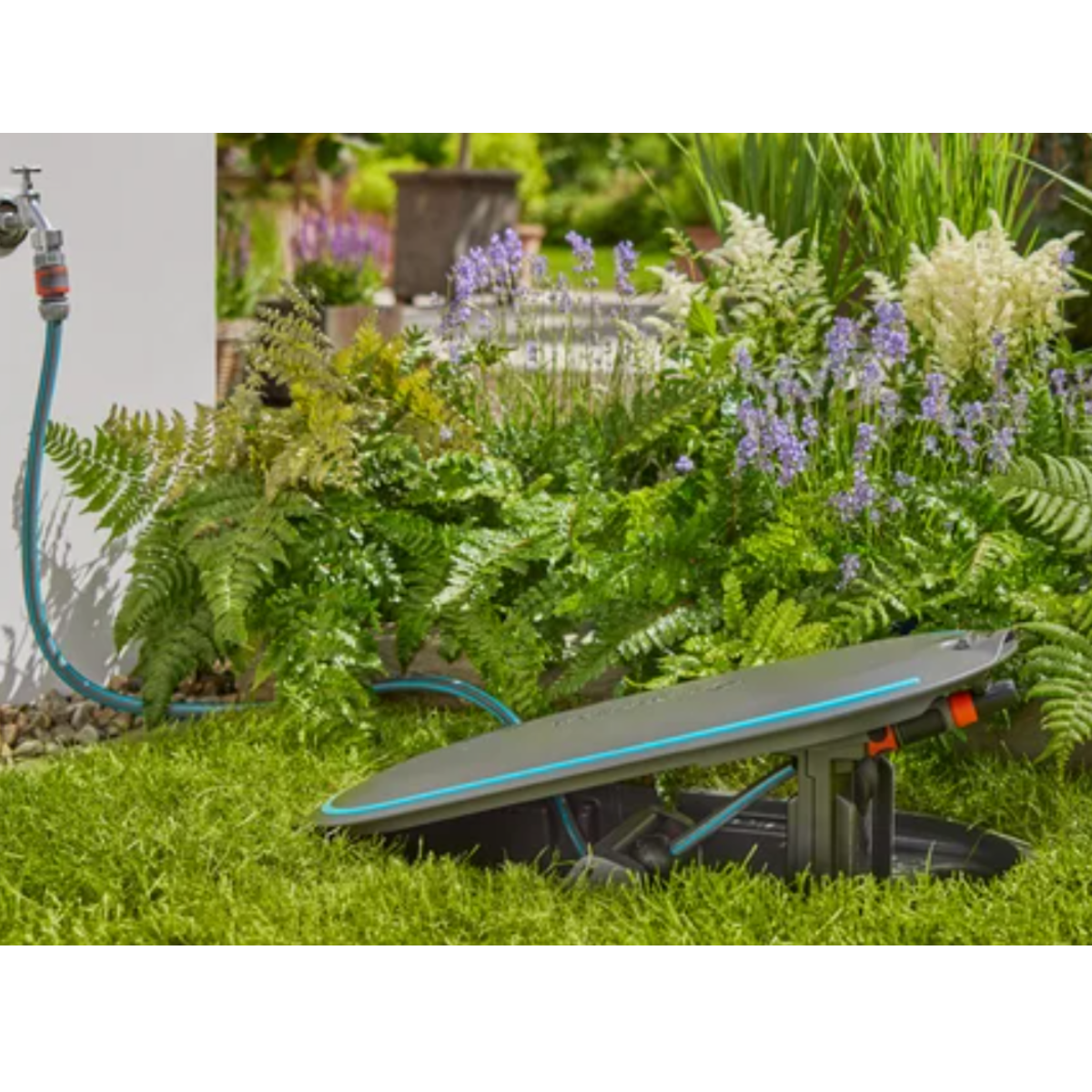 Sistema di irrigazione su erba verde con piante e un muro sullo sfondo.