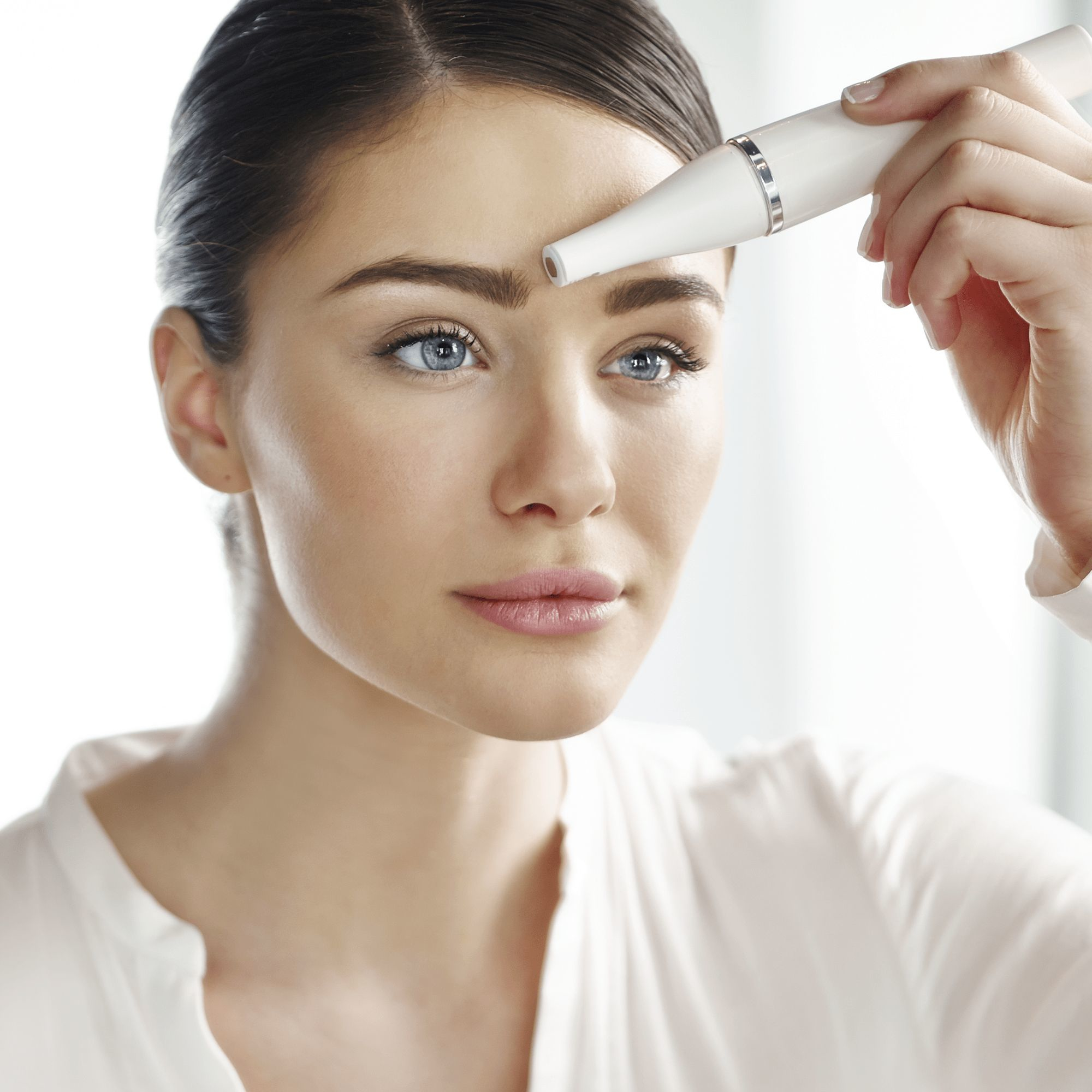 Kobieta używa epilatora do twarzy na swojej brwi. Ma niebieskie oczy i się uśmiecha.