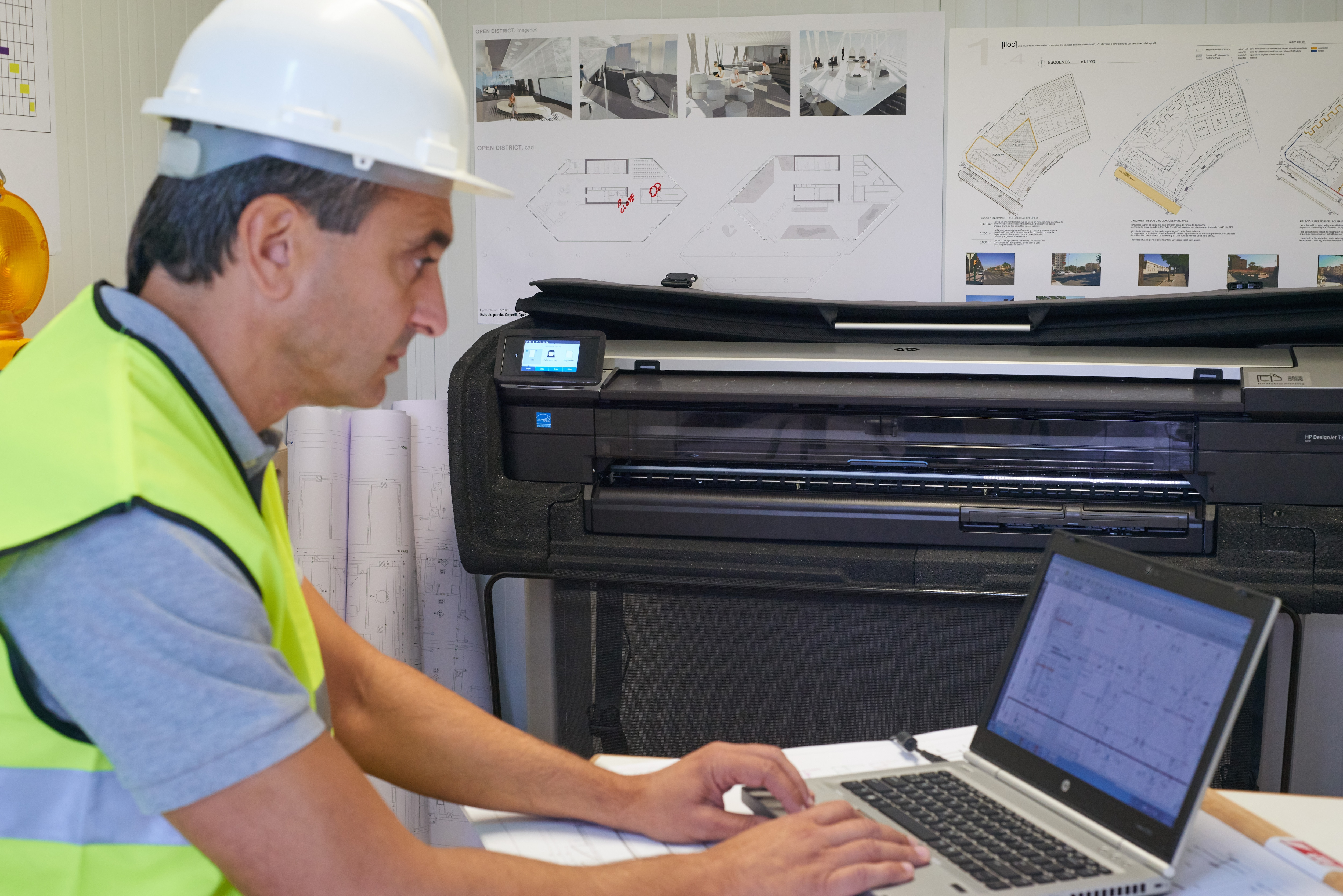Mężczyzna w białym kasku pracuje na laptopie. Drukarka i rysunki w tle.