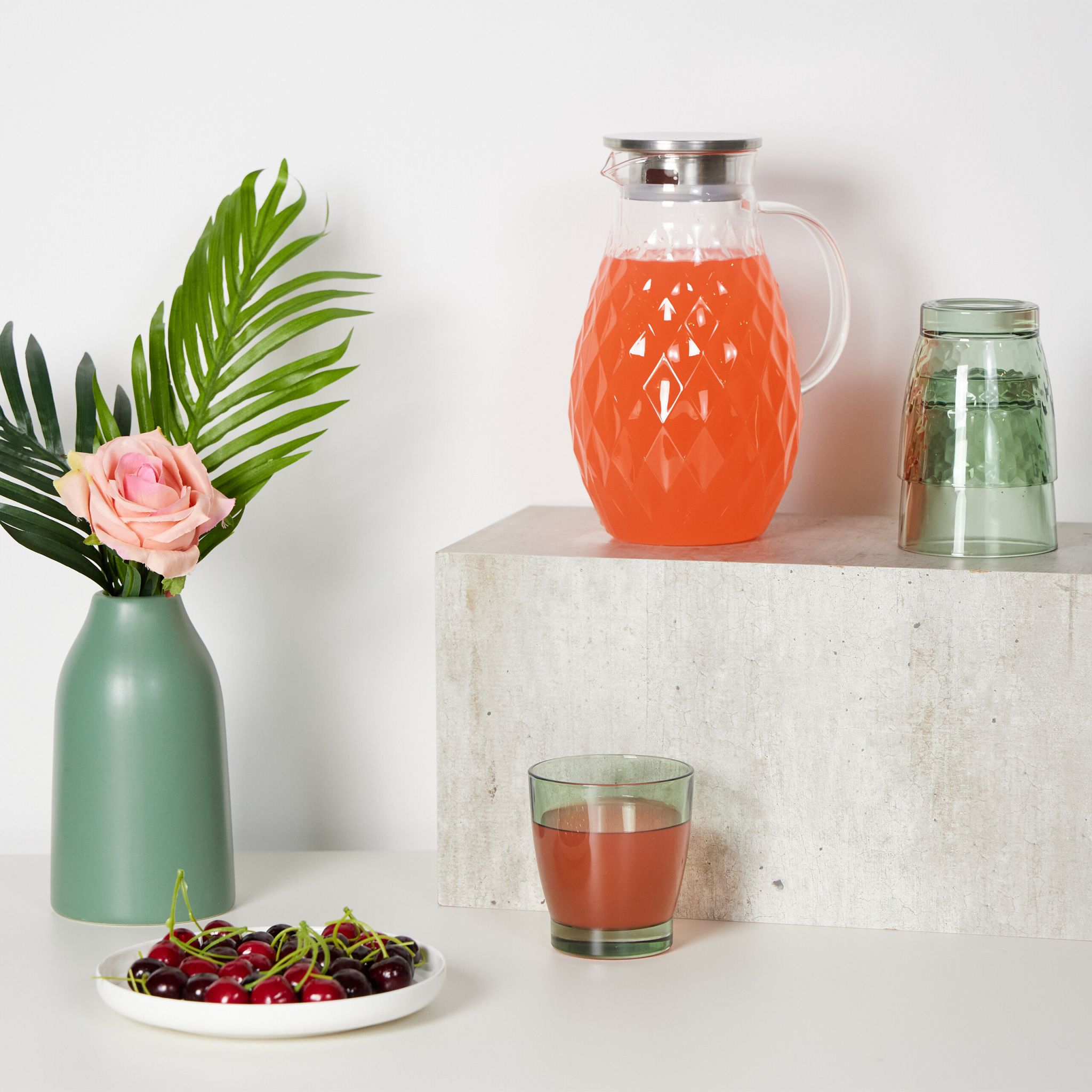 Ein Tisch mit grüner Vase, rosa Blume, Obst und einem Krug.