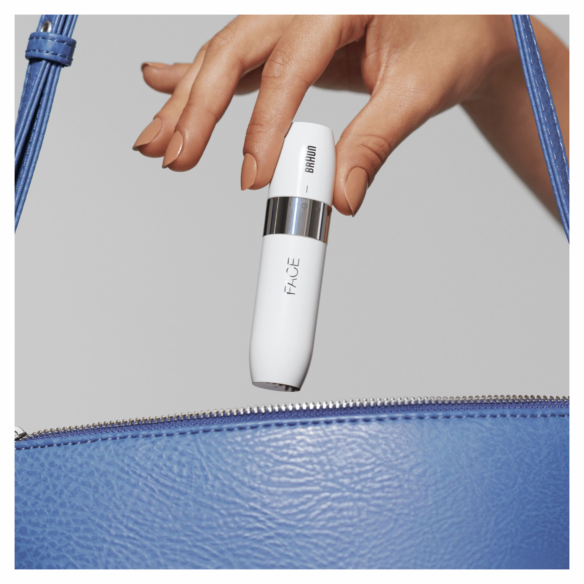 Una mano sosteniendo un dispositivo Braun Face blanco con un bolso azul de fondo.