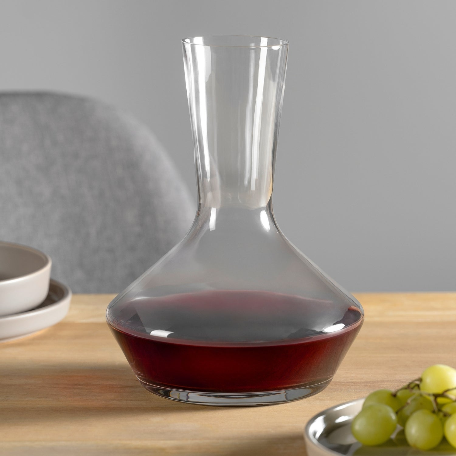 Une carafe à vin en verre transparent avec du vin rouge, des raisins et un fond gris.