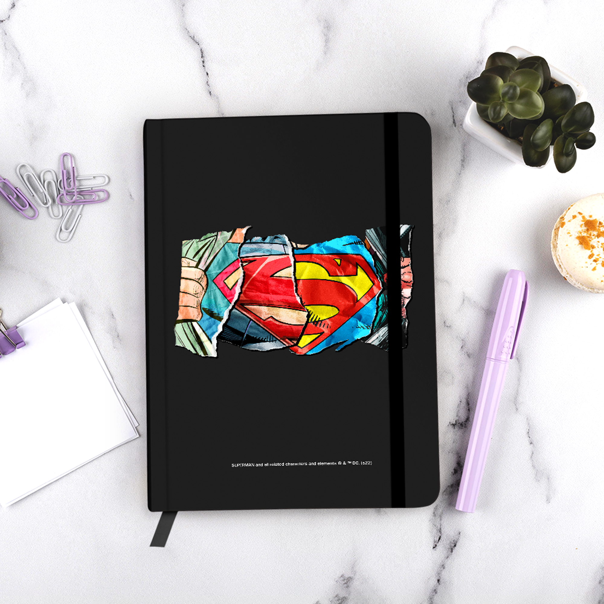 Czarny notatnik z logo Supermana i długopisem na marmurowej powierzchni.