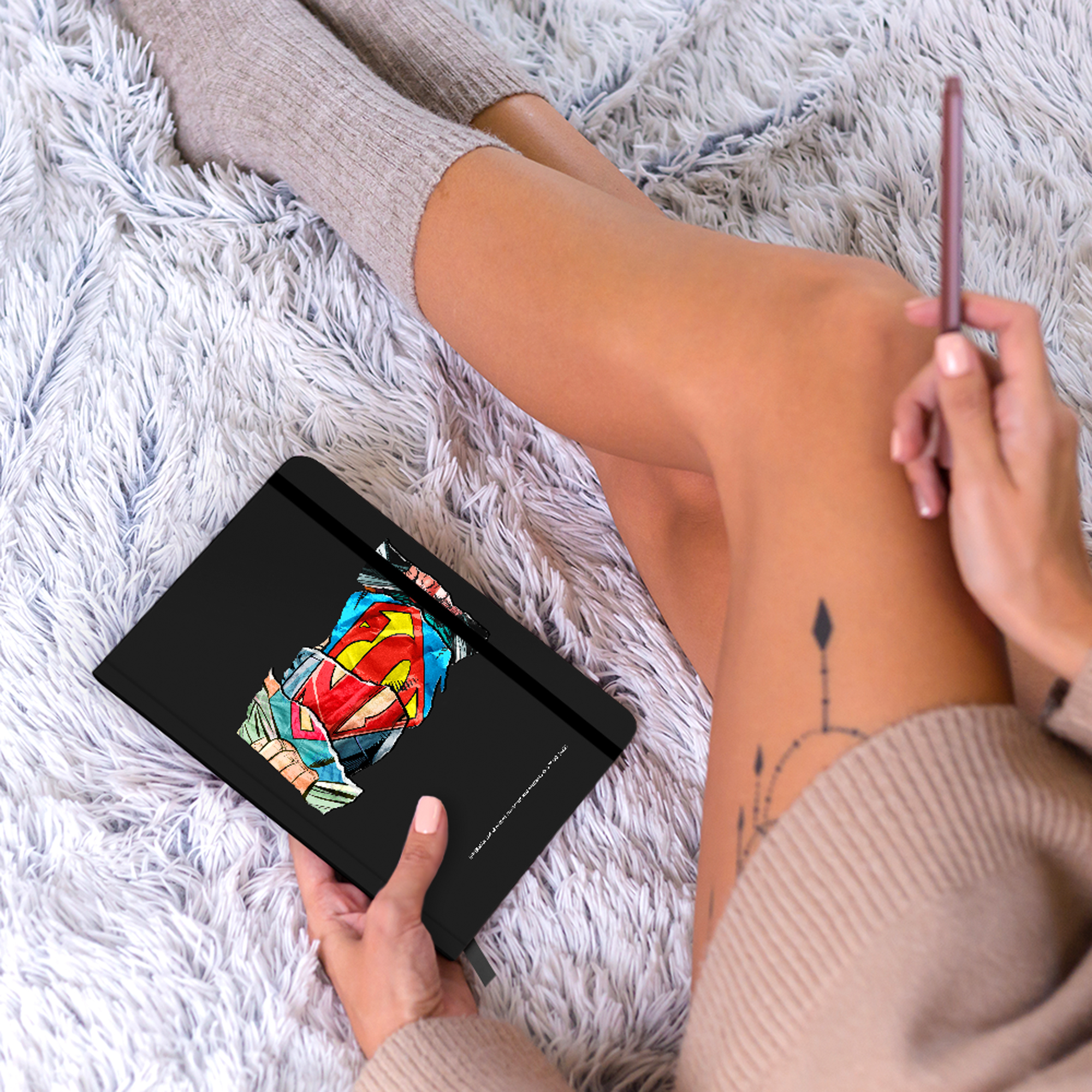 Osoba trzymająca czarny notatnik z nadrukiem Supermana, widoczne nogi i tatuaż.