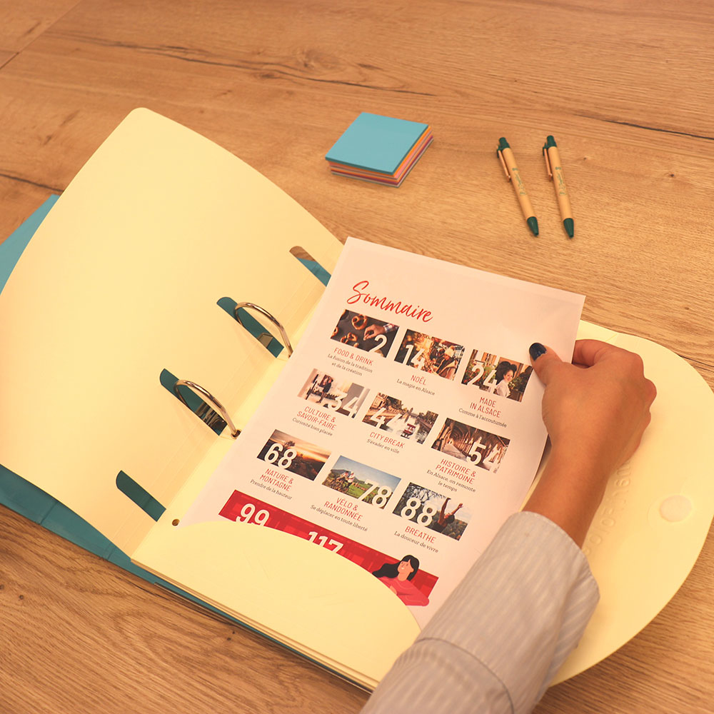 Ręce trzymają broszurę w otwartym segregatorze. Długopisy i karteczki samoprzylepne leżą na drewnianym stole.