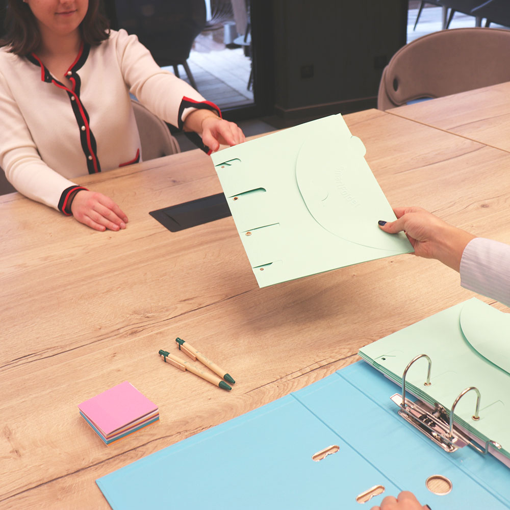 Dwie osoby wymieniające segregator i broszurę na drewnianym stole. Widoczne są długopisy i karteczki samoprzylepne.