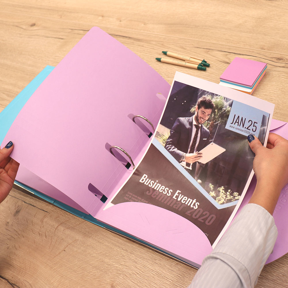 Ręce trzymają broszurę w otwartym segregatorze. Długopisy i karteczki samoprzylepne leżą na drewnianym stole.