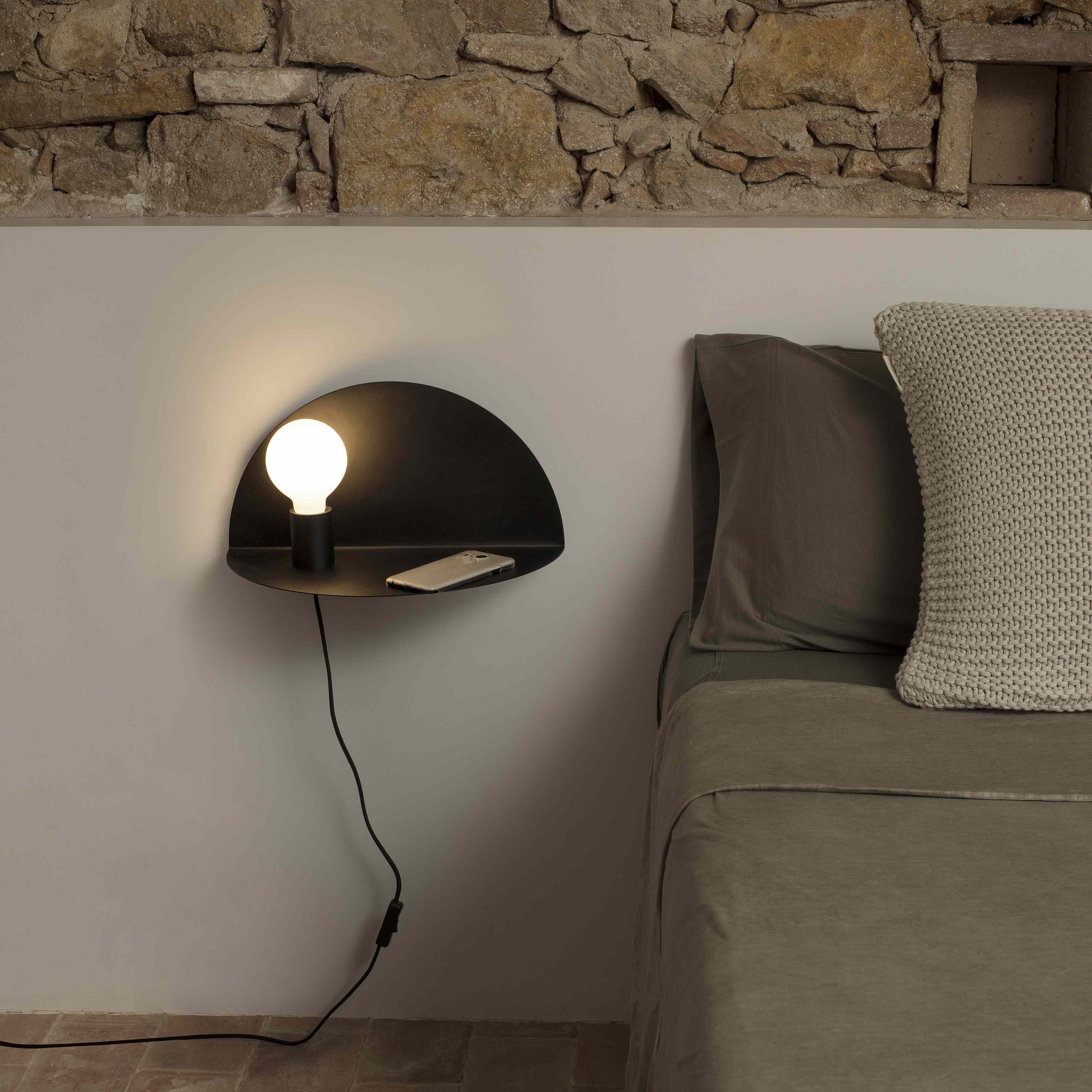 Una lampada da parete nera con una lampadina è su una mensola. Un telefono è appoggiato sulla mensola. Il muro è beige.