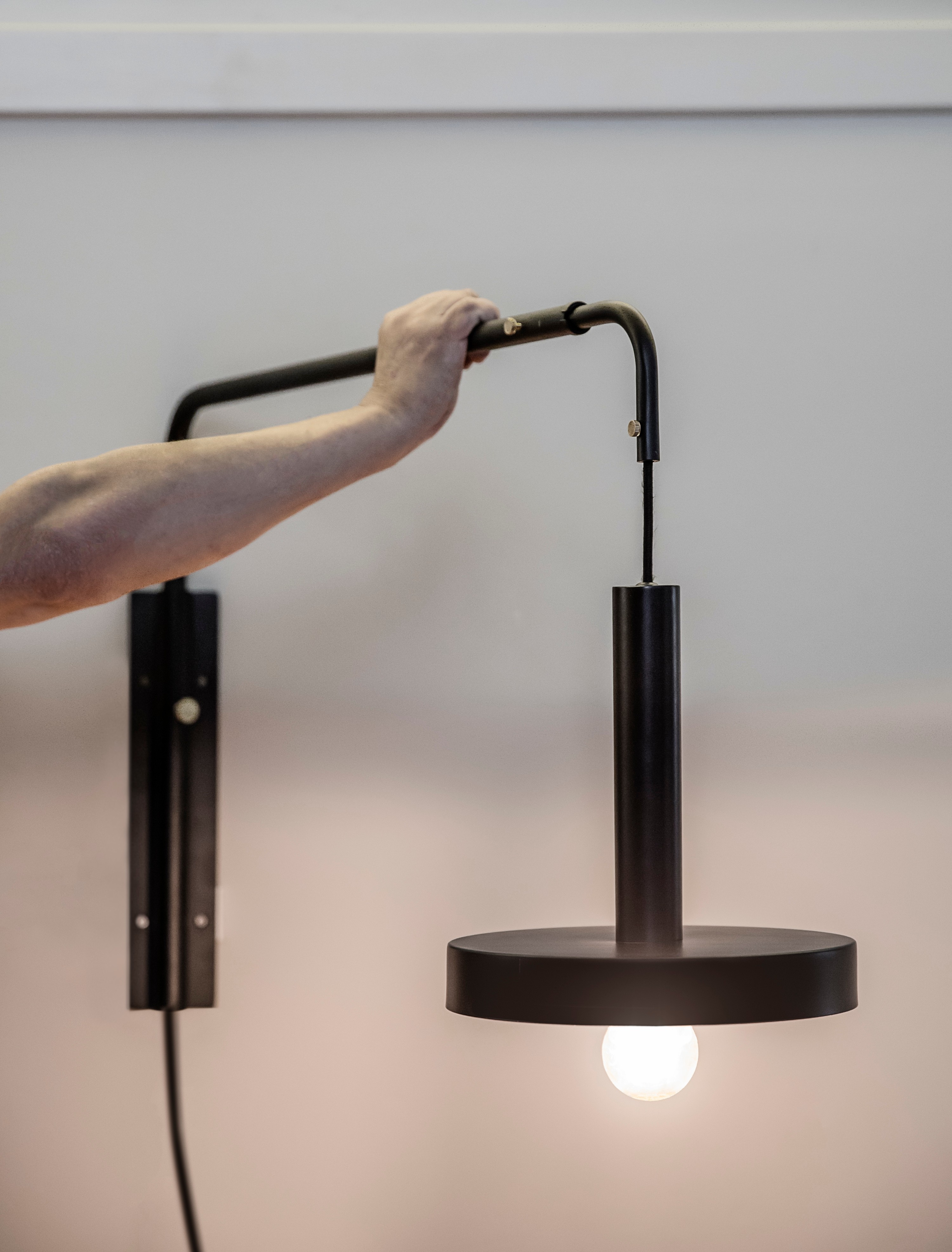 Una mano regola una lampada da parete nera. La lampada ha un corpo cilindrico e un paralume circolare.