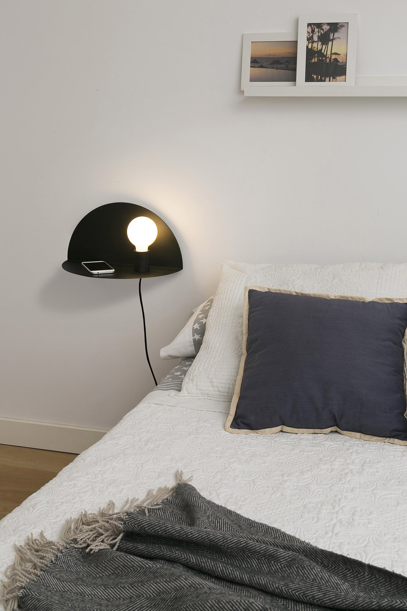 Una lampada da parete nera con lampadina bianca, un telefono e un cuscino grigio su un letto.