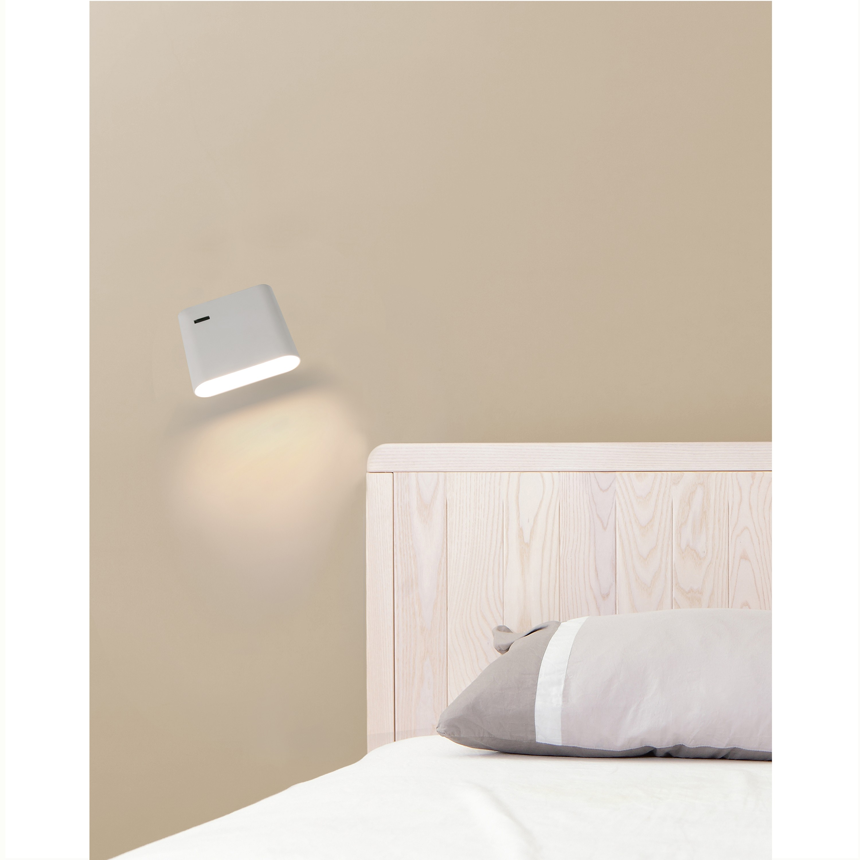 Lampada bianca sulla parete. La lampada è inclinata verso il basso, con un letto bianco e un cuscino in primo piano.
