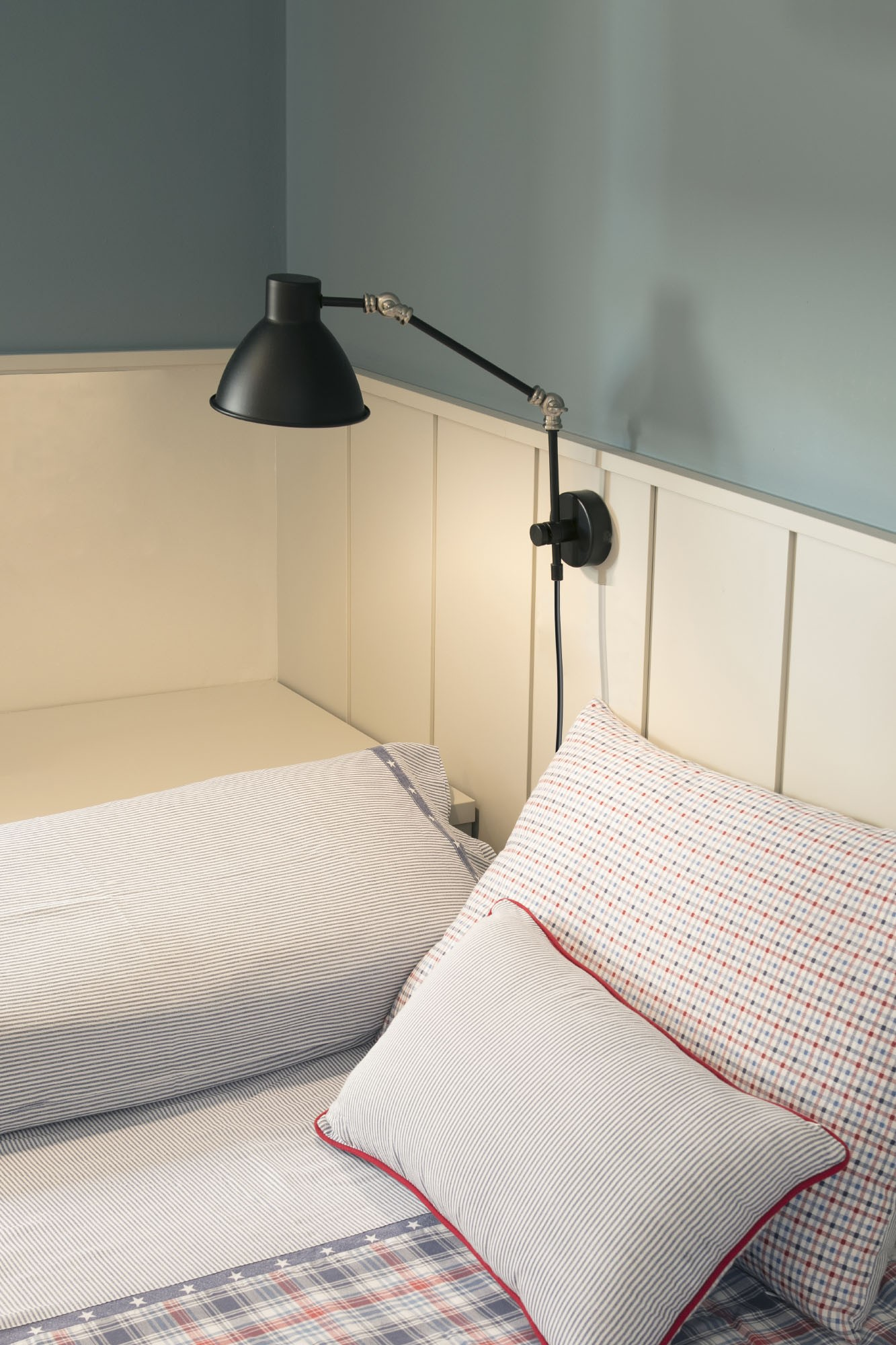 Un letto con cuscini e una lampada nera sul muro. Le pareti sono blu e bianche.