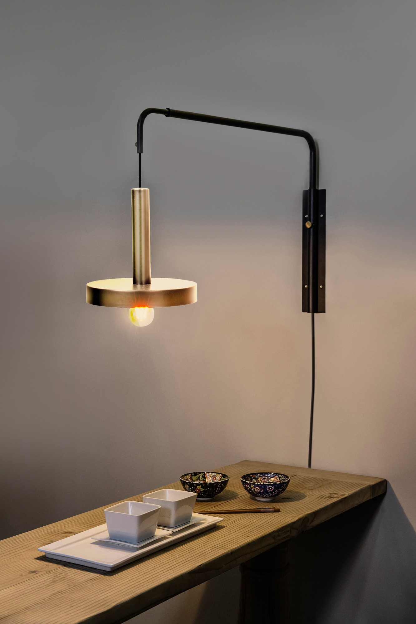 Una lampada da parete con paralume dorato e braccio nero. Un tavolo con ciotole è visibile.