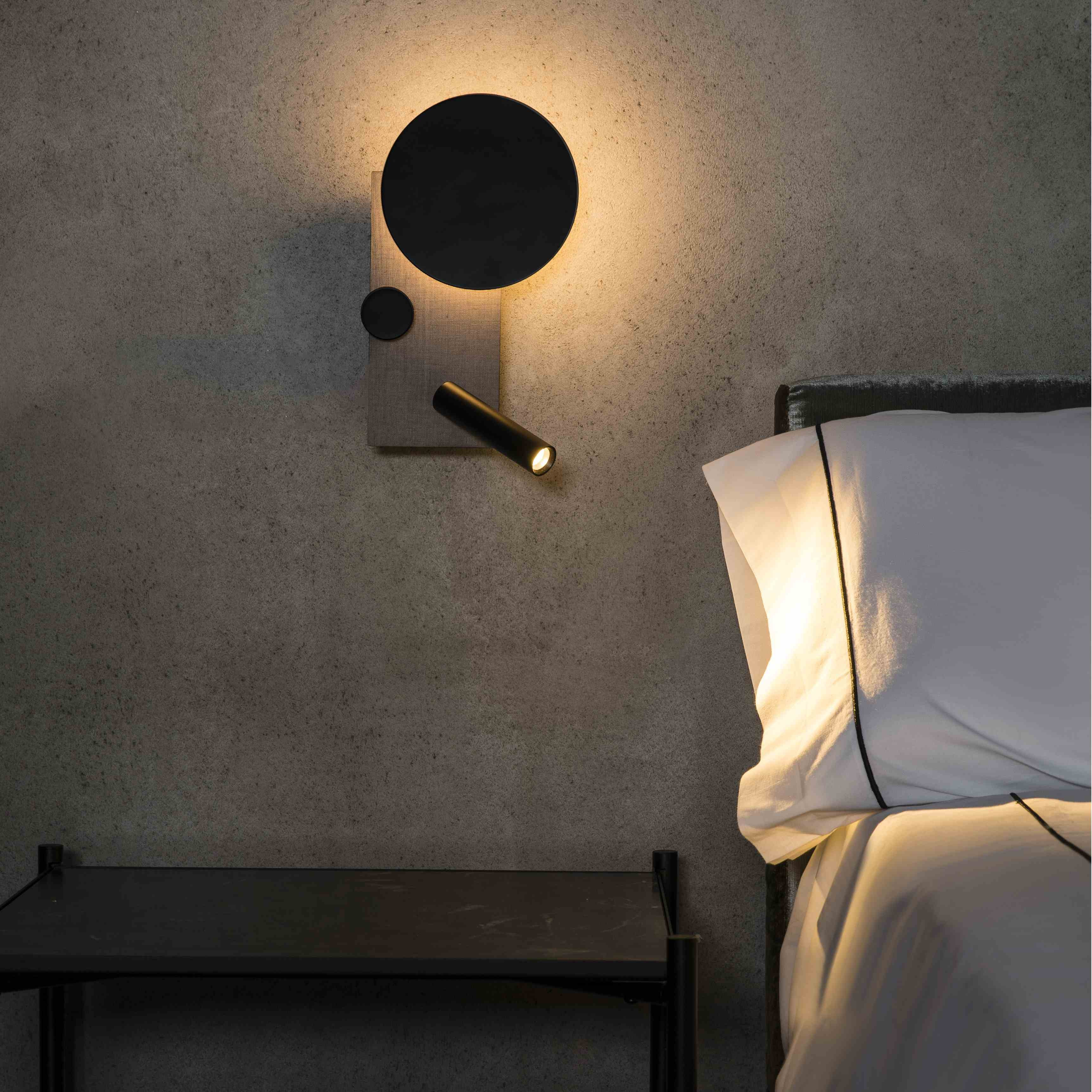 Lampada da parete con luce nera, elemento rotondo e base in legno chiaro. Sono visibili un letto e un comodino.