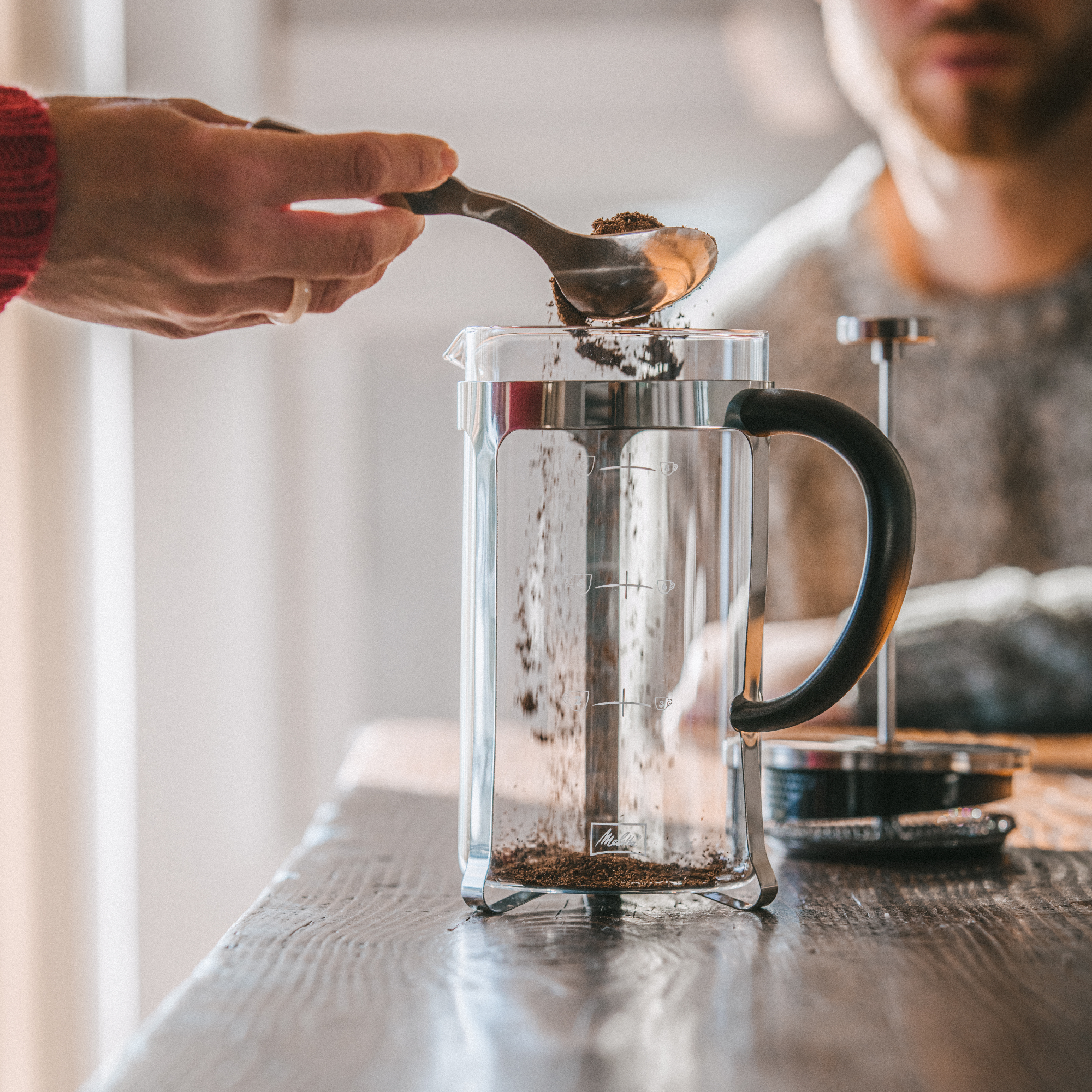Ręka wsypuje zmieloną kawę do prasy francuskiej. Osoba siedzi w tle.