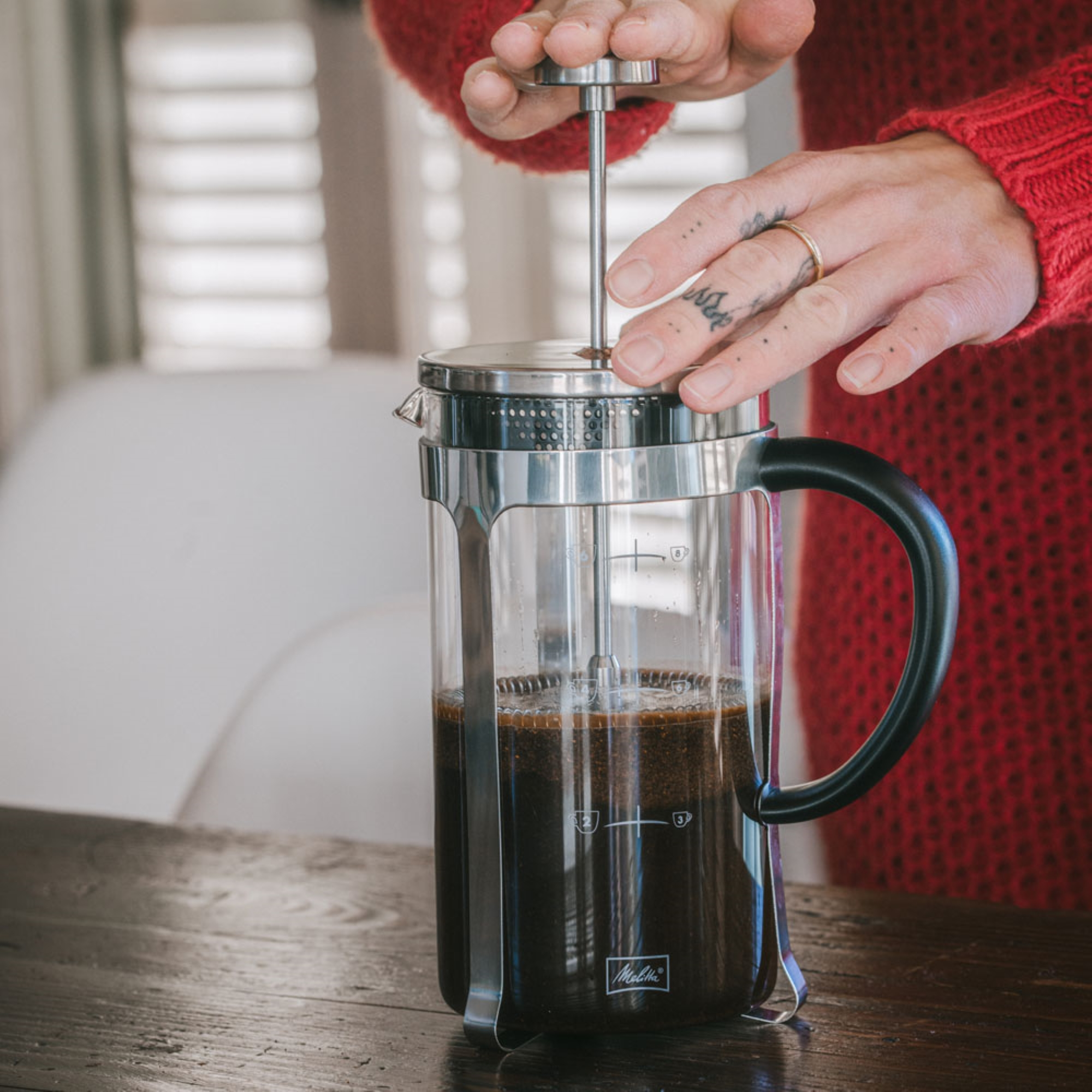 Ręce wciskają kawę w szklany french press. Czerwony sweter w tle.