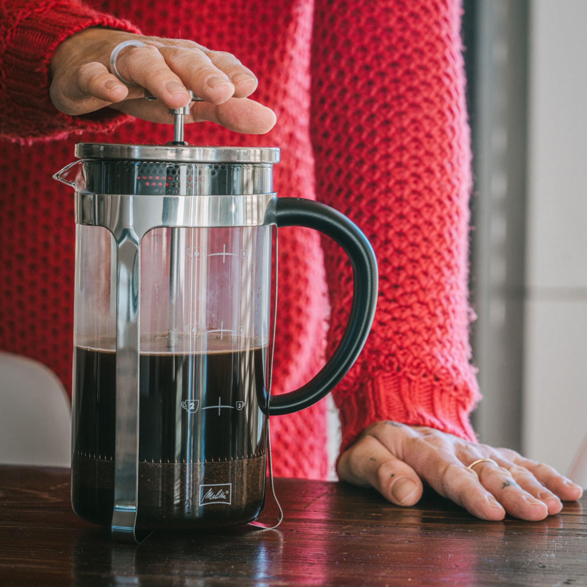 Osoba używająca kawiarki french press. Czerwony sweter i drewniany stół.