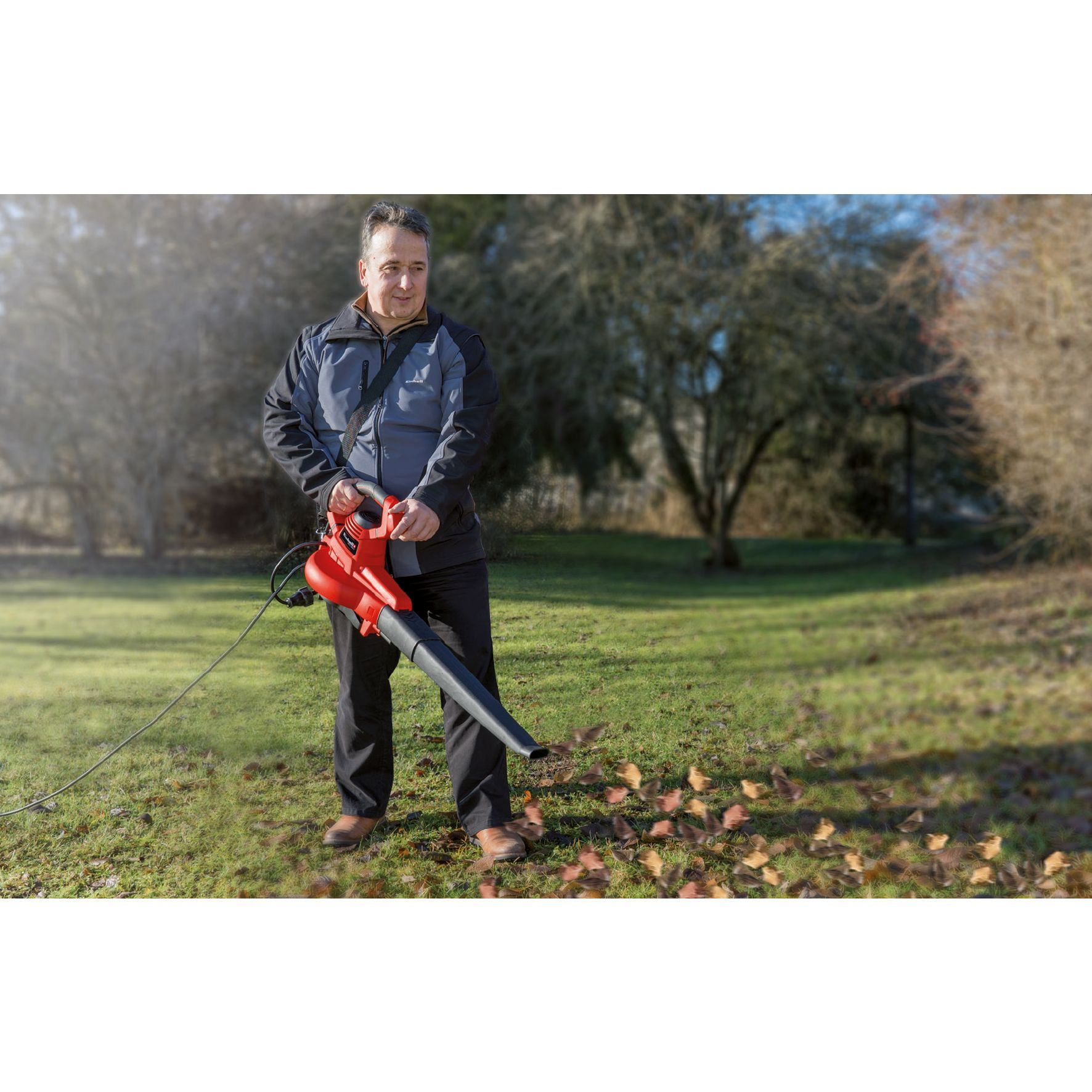 A man uses a red leaf blower on a grassy area with fallen leaves, trees in the background, and a blue sky.