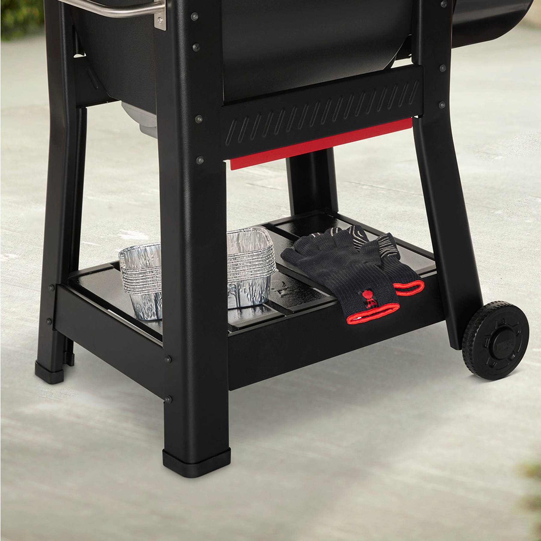 Parrilla negra con detalles rojos, bandejas de aluminio y guantes en el estante. Rueda a la derecha.