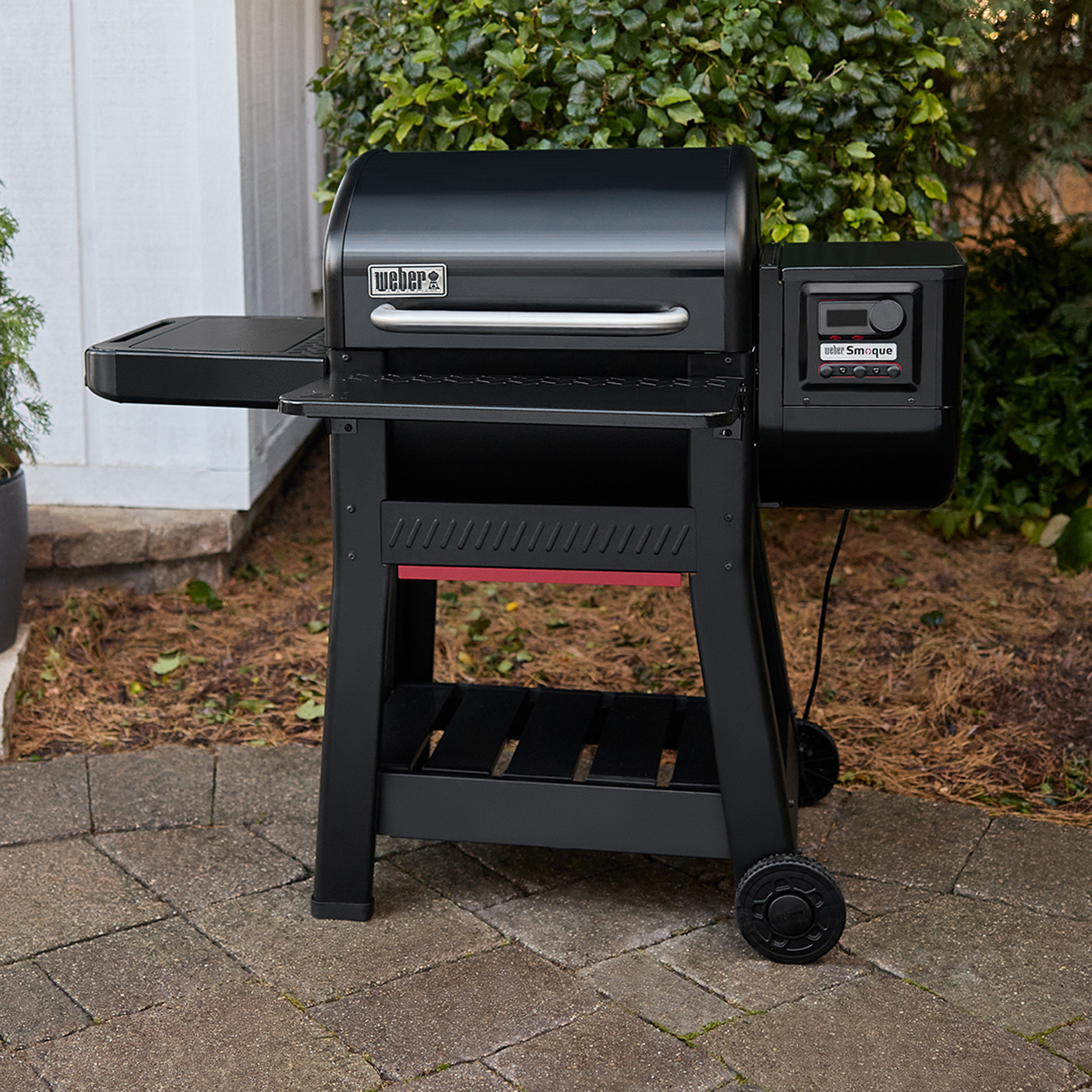 Parrilla Weber negra en un patio, con mesa lateral y ruedas, al aire libre.