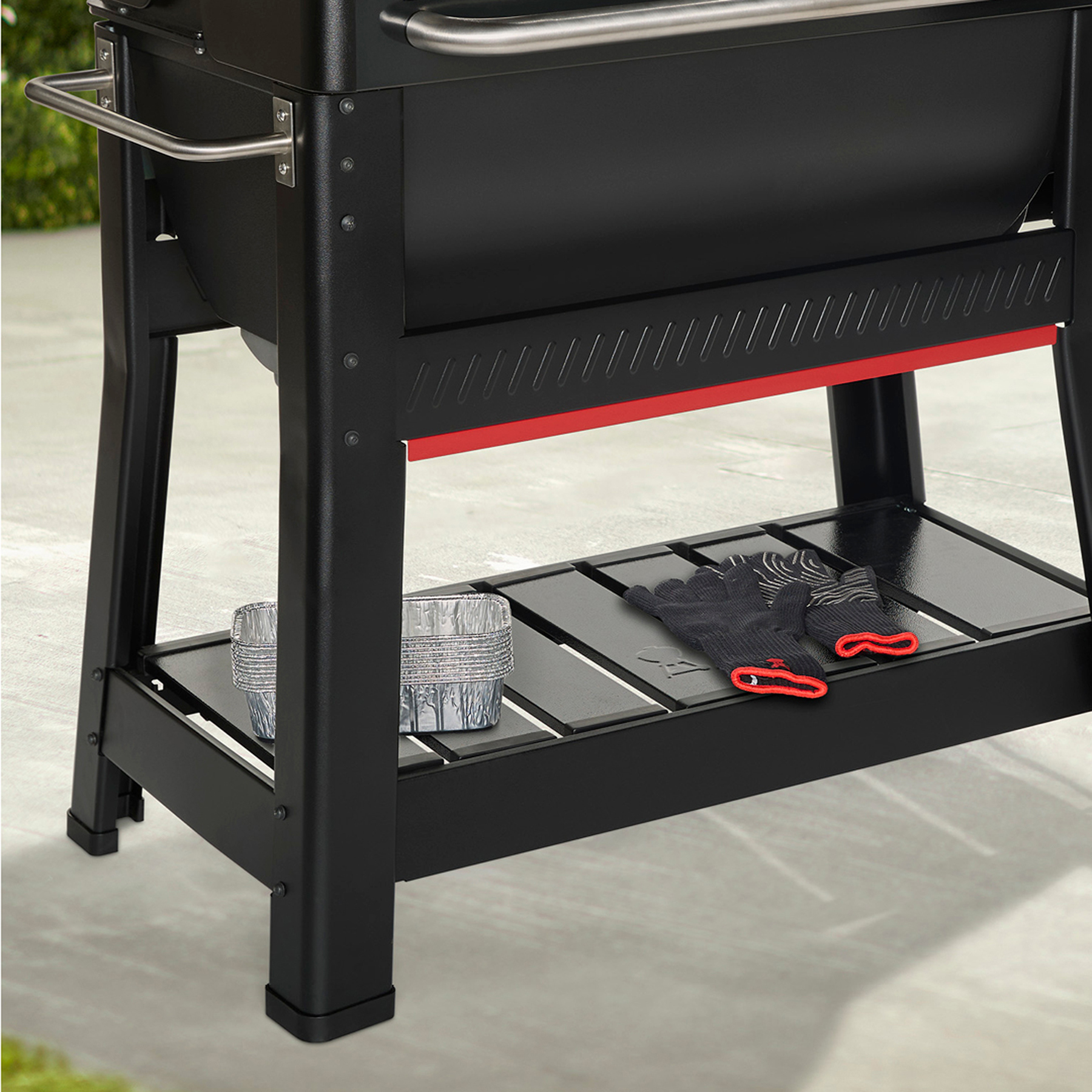 Griglia Weber con ripiano, contenitori in alluminio e guanti. Accenti rossi. Sfondo bianco.