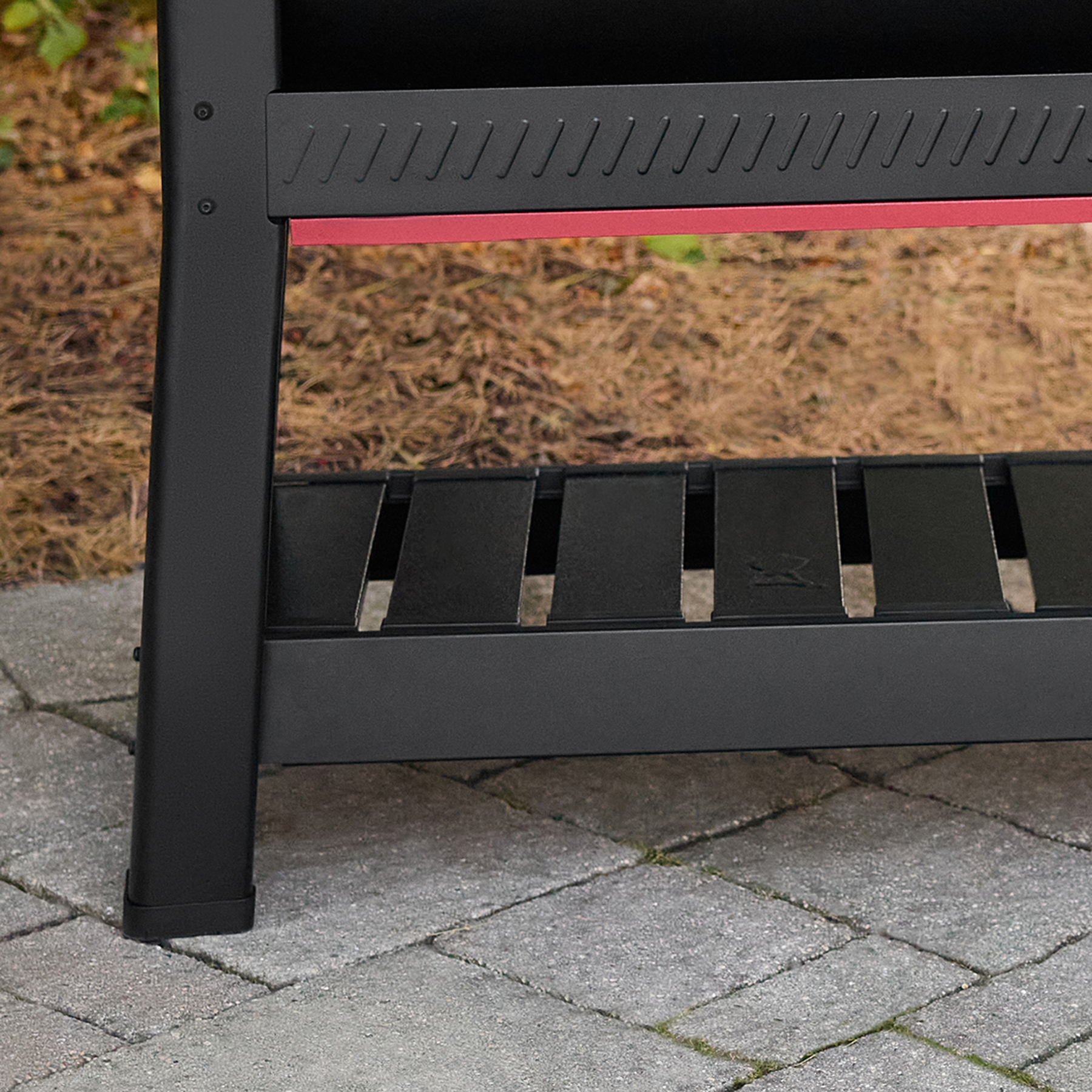 Struttura nera del grill con accento rosso, su pavimentazione grigia, erba sullo sfondo.