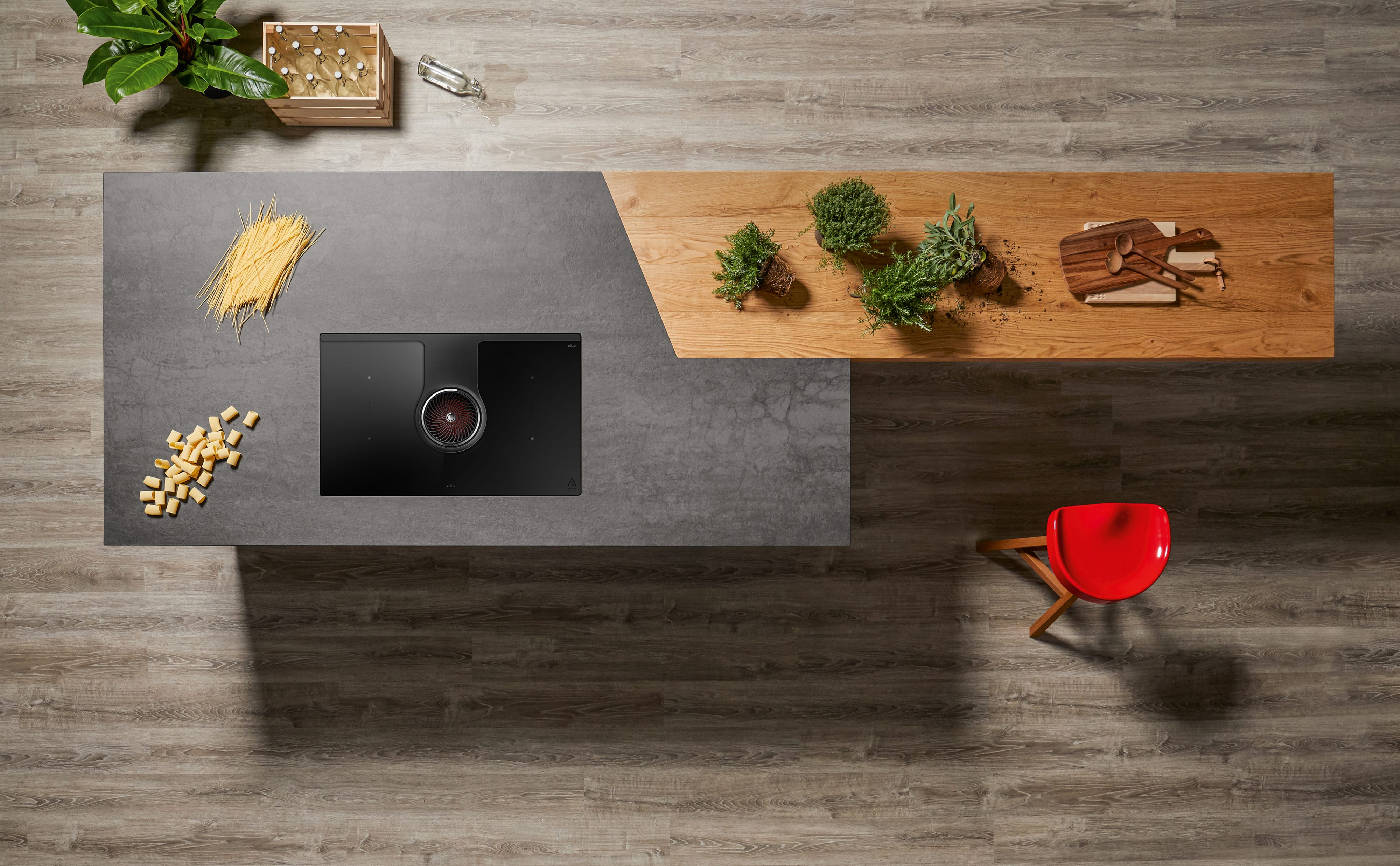 Isola cucina con piano cottura, cibo e sedia rossa su pavimento in legno. Piante verdi e una scatola sull'isola.