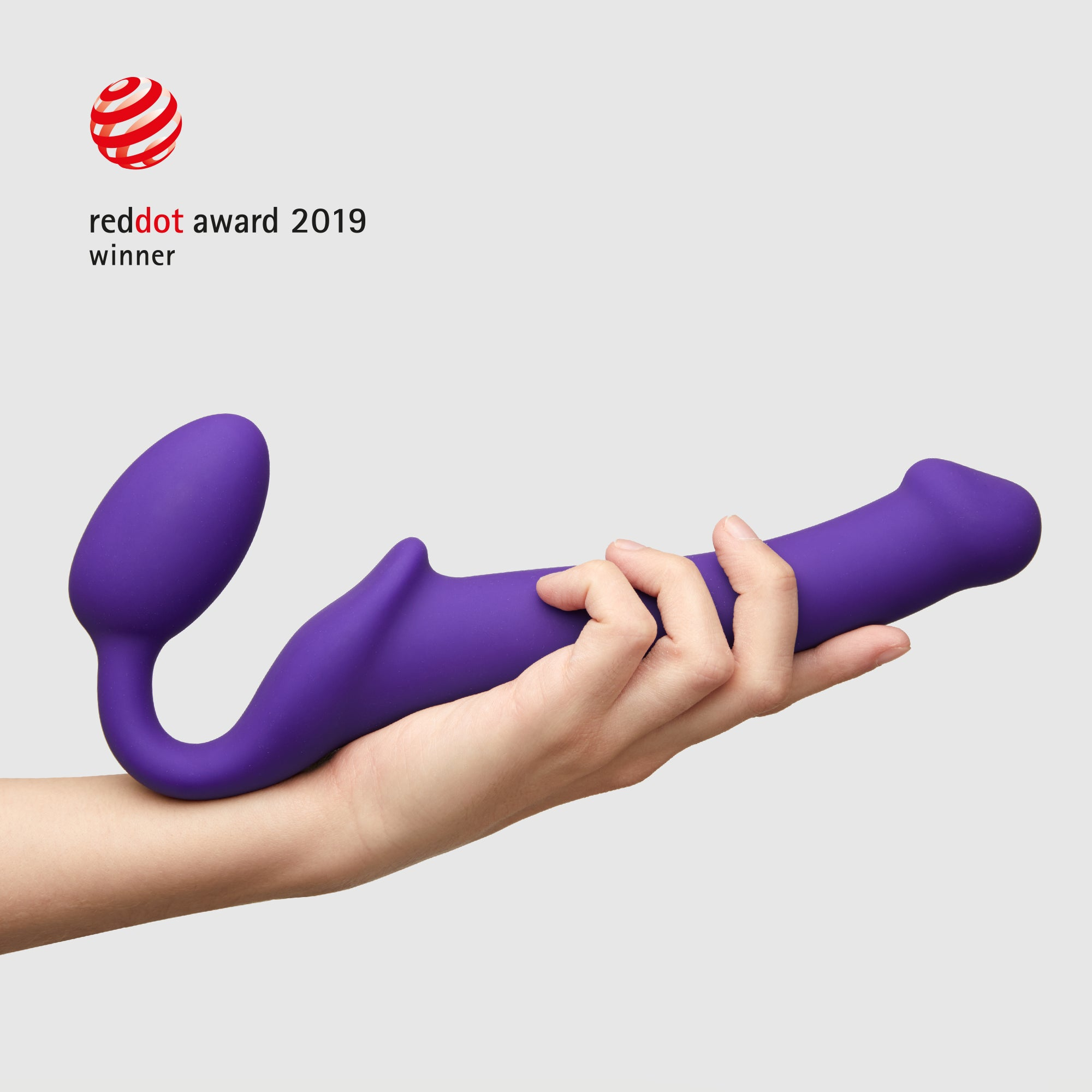 Fioletowa zabawka erotyczna trzymana w dłoni. W tle widać nagrodę Red Dot Award 2019.